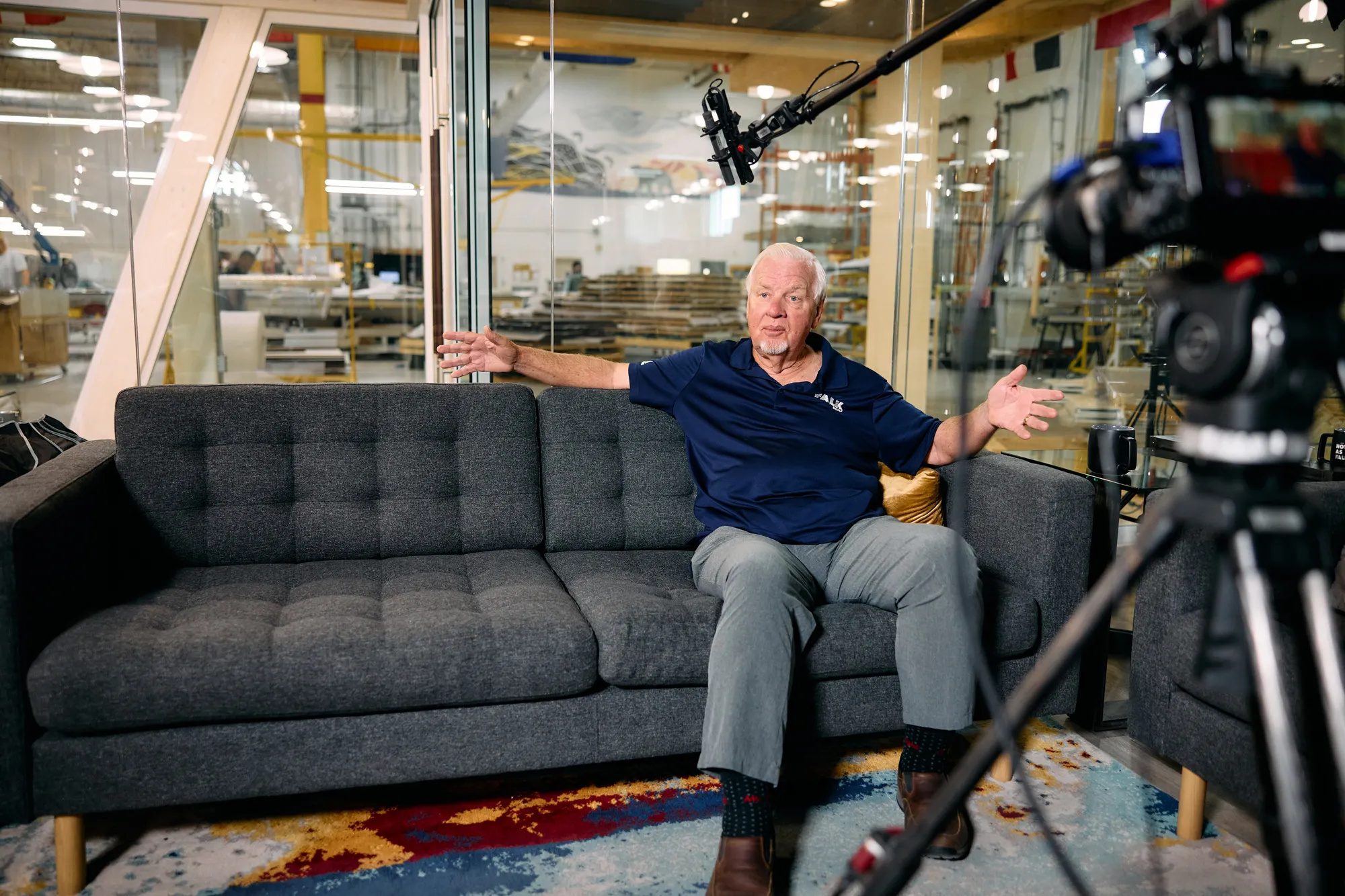 a man sits on a couch in front of a window into a warehouse, he speaks to the camera