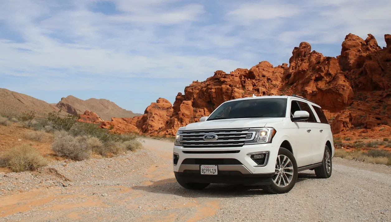 A white SUV drives on a gravel road through a desert landscape with red rock formations.