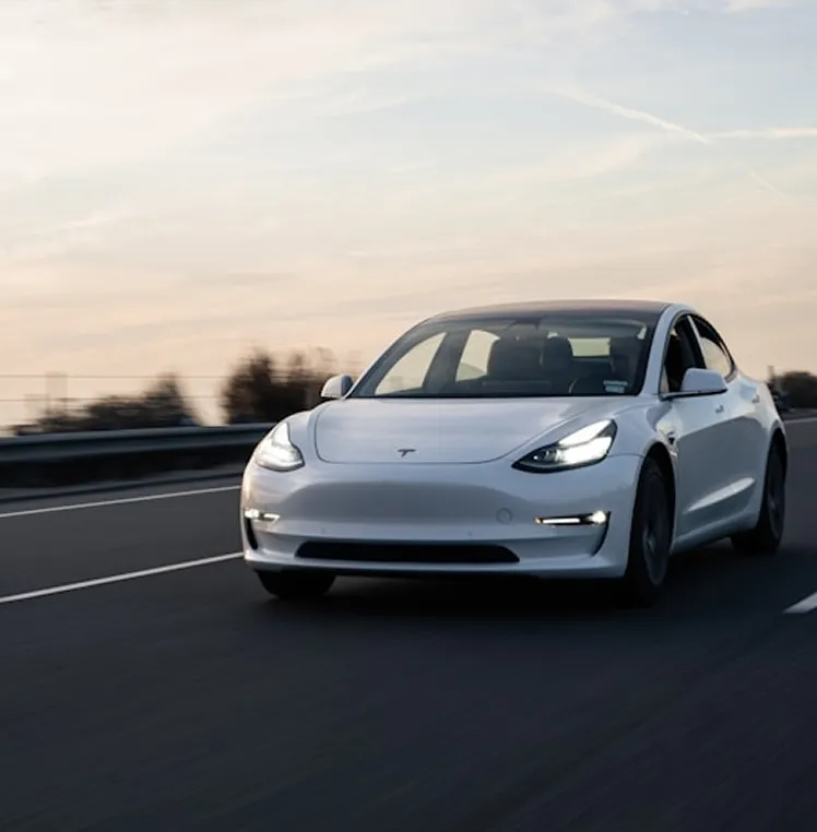 A white Tesla Model 3 driving on a highway at dusk.