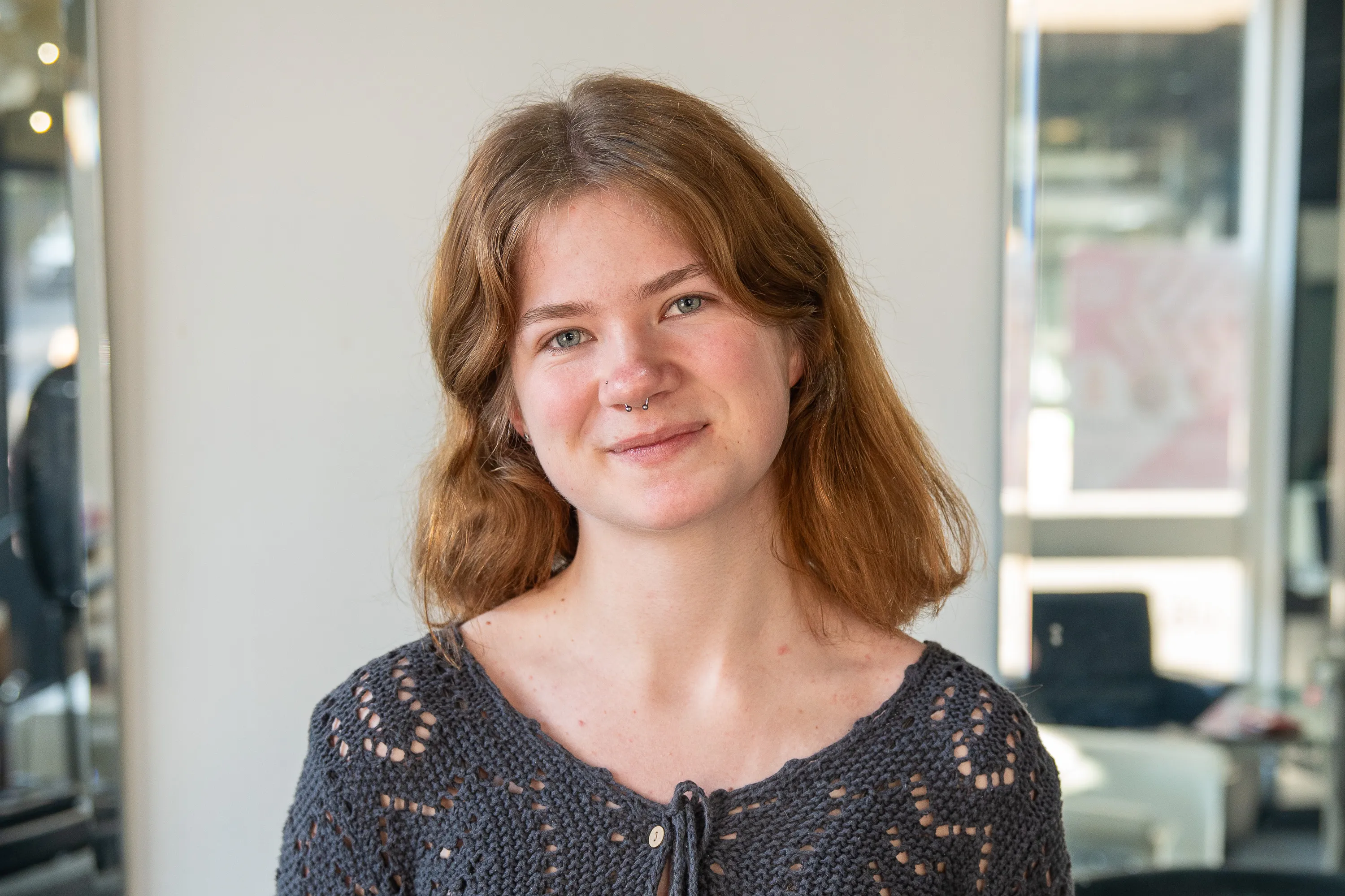 Young woman with light brown hair and septum piercing smiling softly, wearing a dark blue crocheted top.