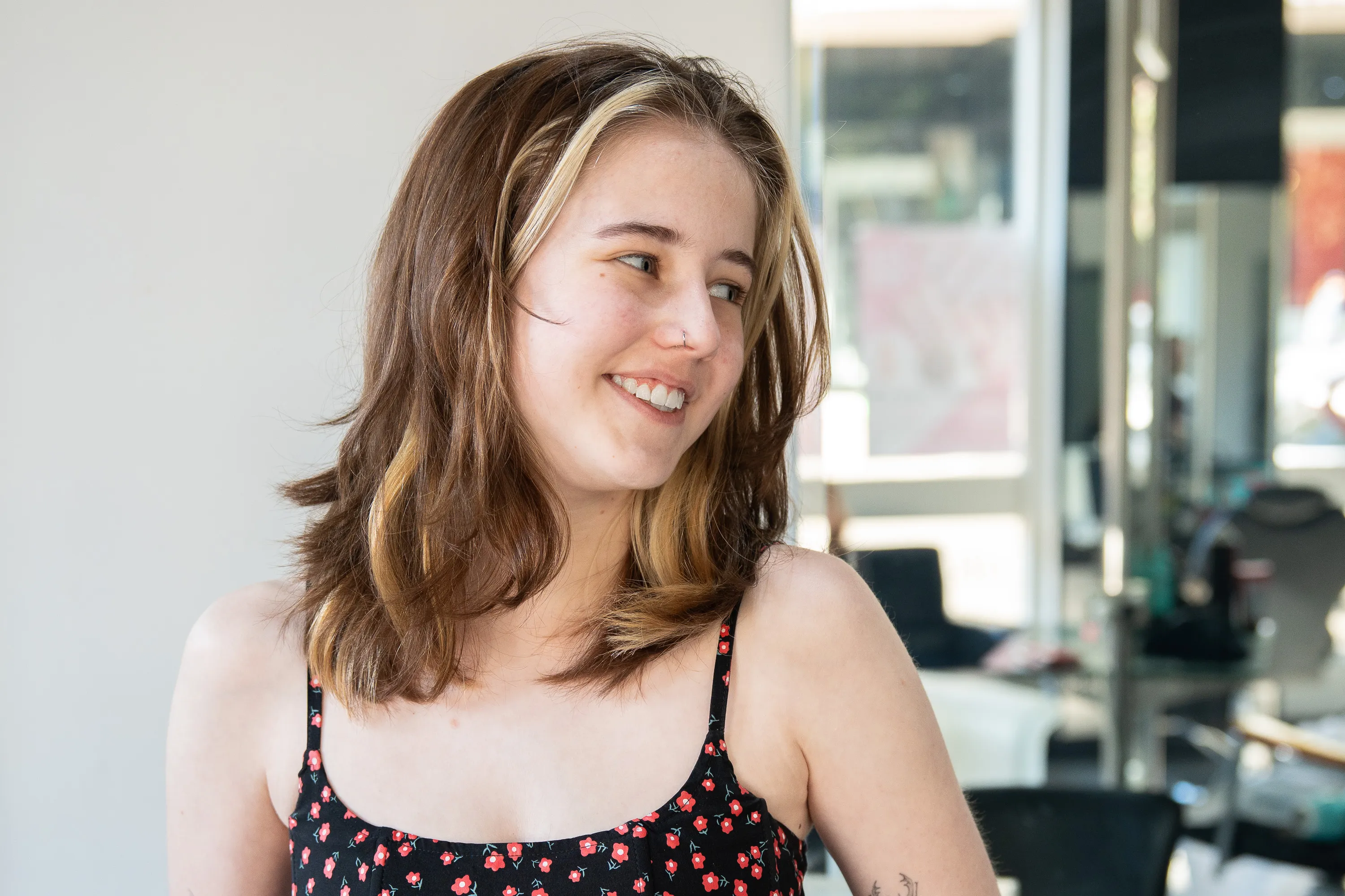 Smiling young woman with shoulder-length brown and blonde hair wearing a black dress with red flowers, looking to her right.