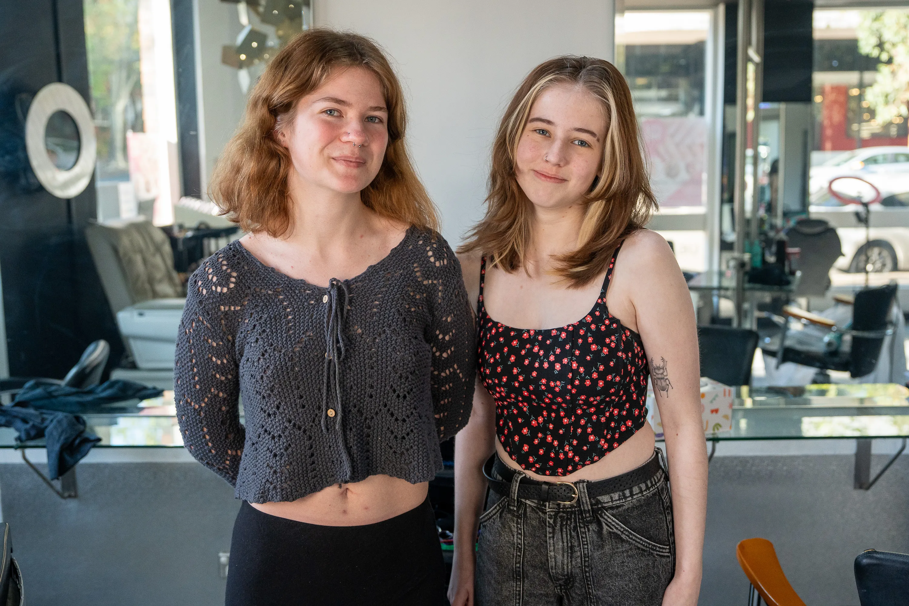 Two young women standing side by side indoors, one with a grey knitted cropped sweater and the other in a black floral sleeveless top with jeans.
