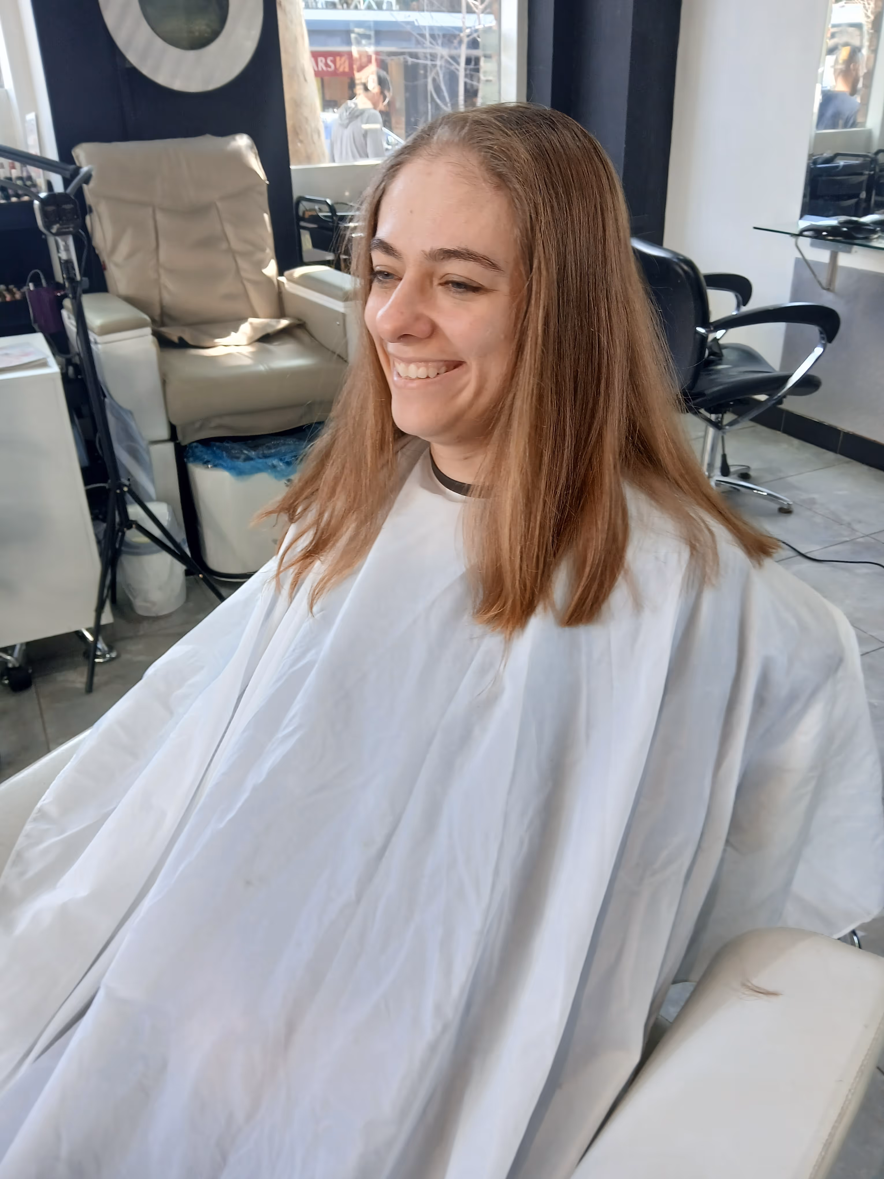 Smiling woman with straight, shoulder-length light brown hair wearing a white salon cape seated in a hair salon.