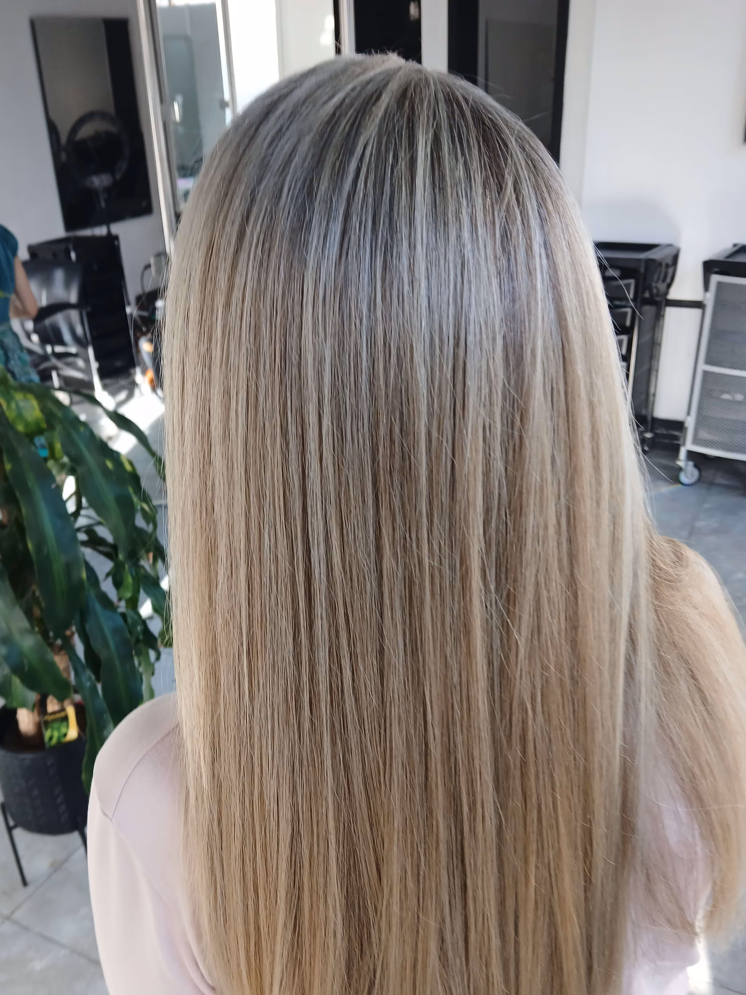 Back view of a person with straight, blonde hair in a salon setting with a plant and salon chairs in the background.