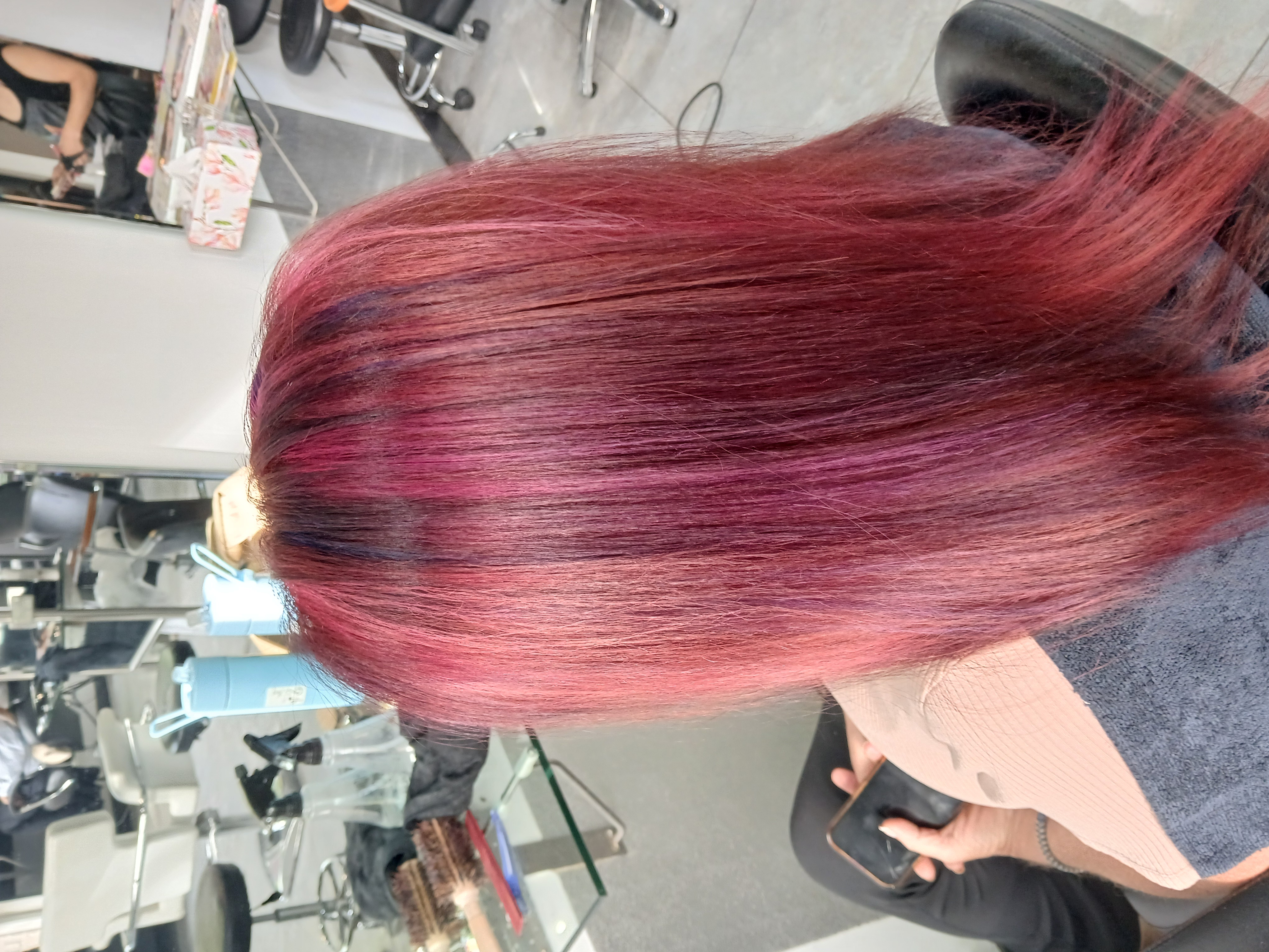 Person with straight, shoulder-length hair dyed in shades of pink and purple sitting in a salon chair holding a phone.