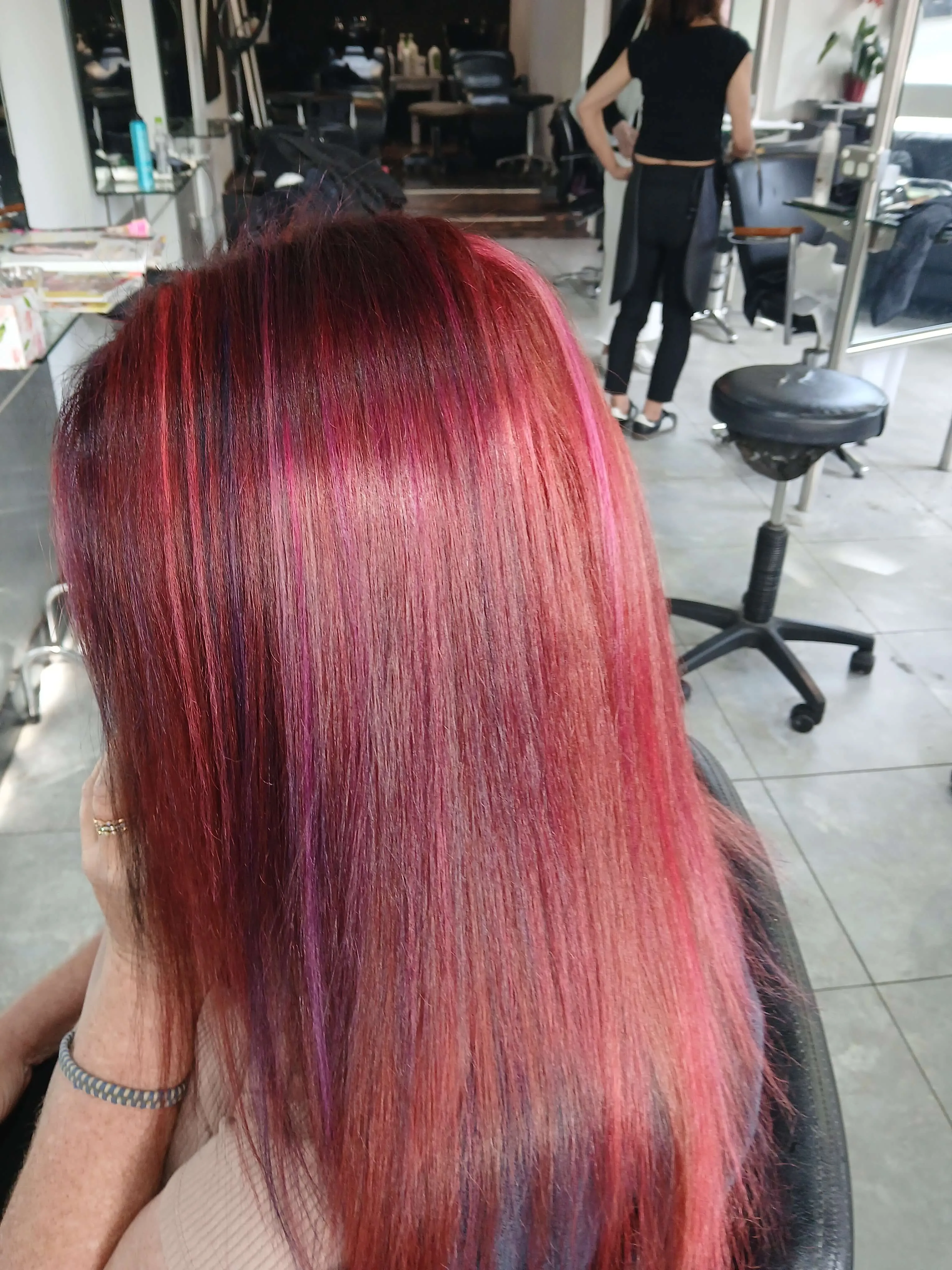 Person with long, straight hair dyed in shades of red, purple, and pink sitting in a salon chair.