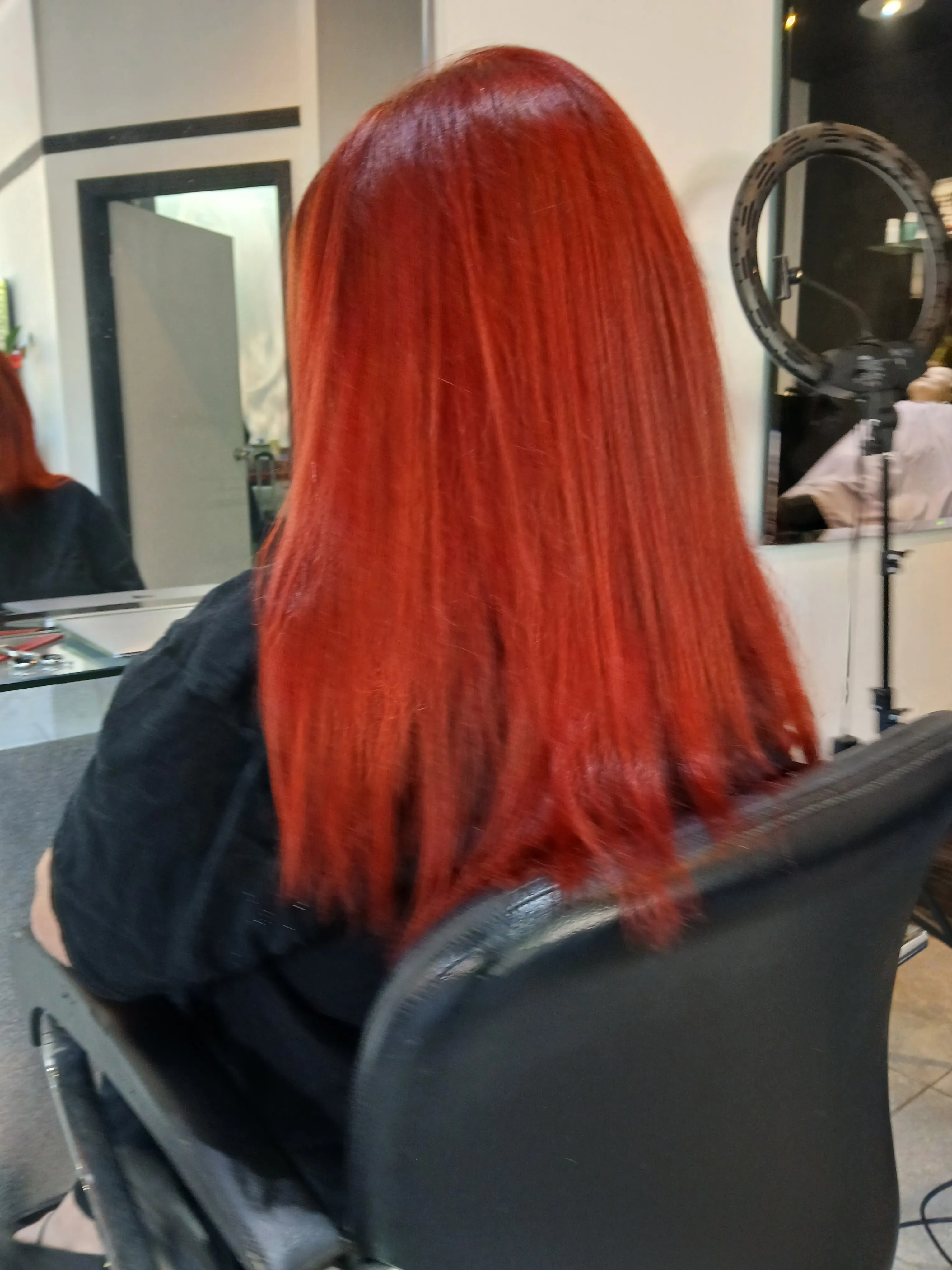 Person with straight, vibrant red shoulder-length hair sitting in a salon chair facing a mirror.