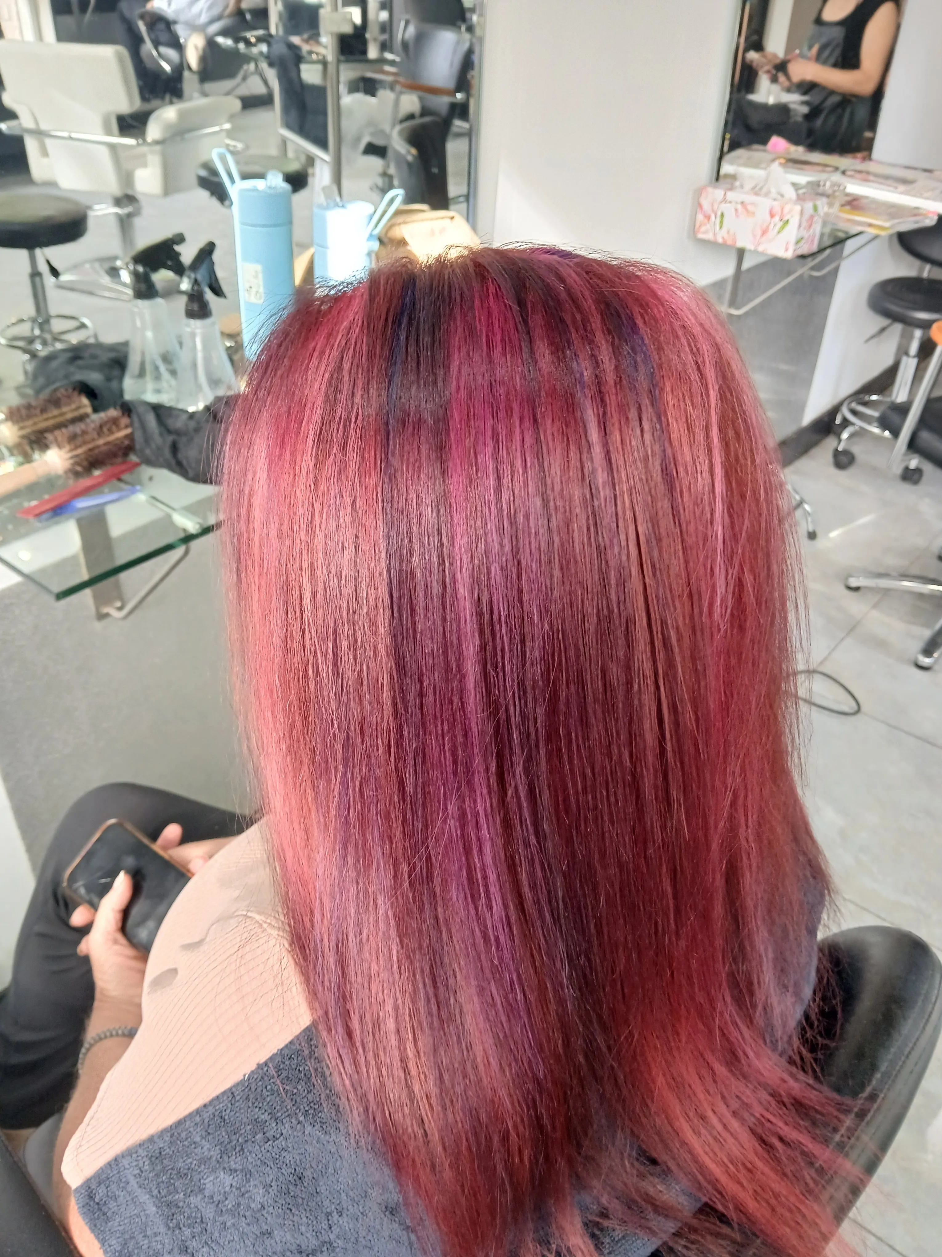 Back view of a person with straight, freshly dyed vibrant pink and purple hair sitting in a salon chair, holding a phone.