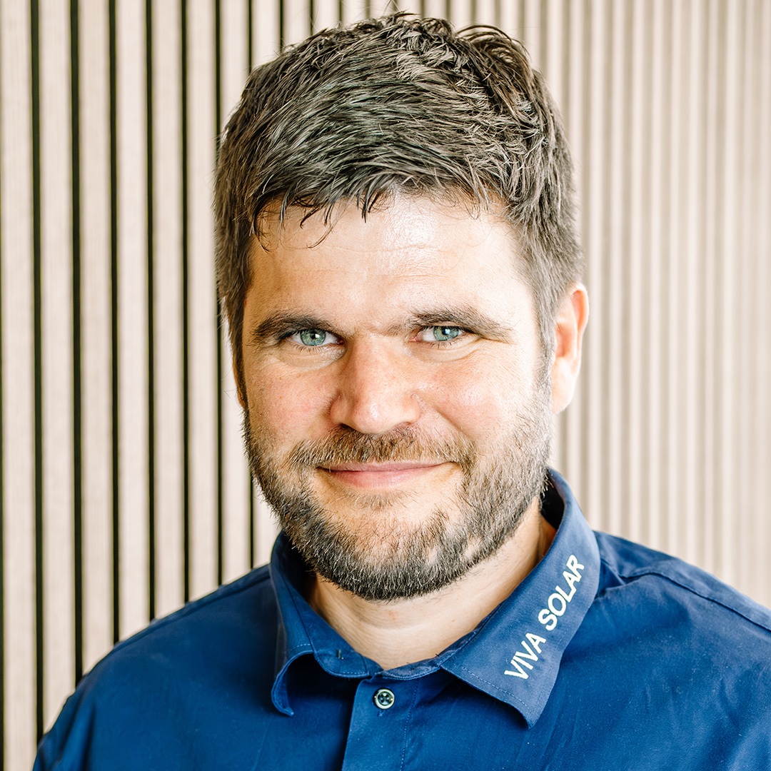 Ein Mann mit kurzen, grauen Haaren und einem freundlichen Gesichtsausdruck trägt ein blaues Hemd. Im Hintergrund sieht man eine Wand mit vertikalen Holzlatten.