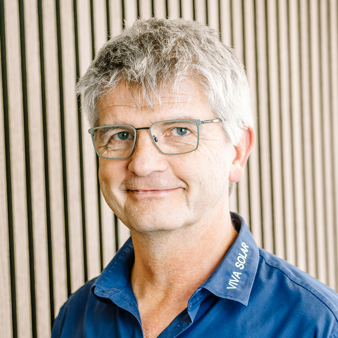 Ein lächelnder Mann mit Brille und grauen Haaren sitzt vor einer modernen Holzpaneelwand. Er trägt ein blaues Hemd und hat eine freundliche Ausstrahlung.