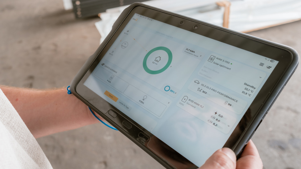 Eine Person hält ein Tablet in der Hand, auf dem eine App mit grafischen Daten und einer Fortschrittsanzeige angezeigt wird. Der Hintergrund zeigt eine unaufgeräumte Fläche.