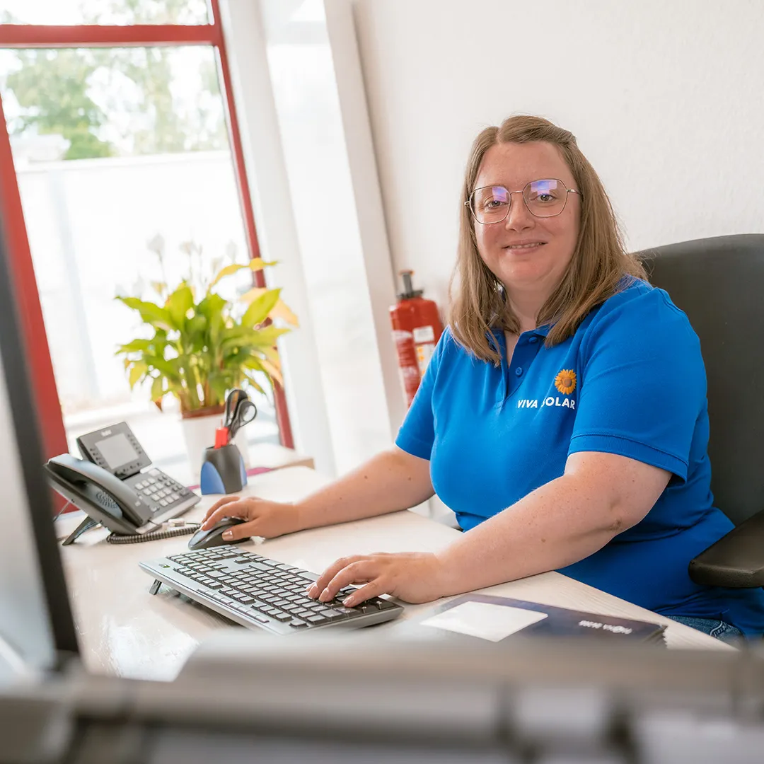 Eine Frau sitzt an einem Schreibtisch in einem hellen, modernen Büro. Sie trägt ein blaues Polo-Shirt und lächelt freundlich, während sie an einem Computer arbeitet. Im Hintergrund ist eine grüne Pflanze und ein Feuerlöscher sichtbar.