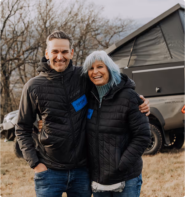 Ein lächelndes Paar in schwarzen Jacken steht eng umarmt im Freien vor einem Zelt auf einem Geländewagen.