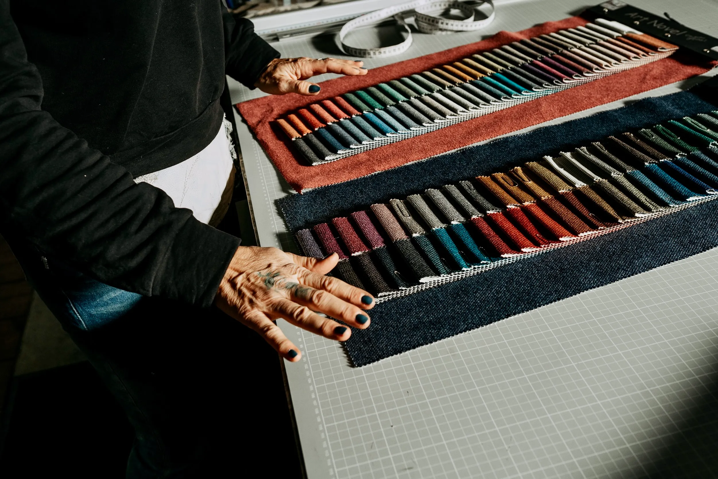 Person mit tätowierten Händen betrachtet Stoffmuster in verschiedenen Farben auf einem Schneidetisch.
