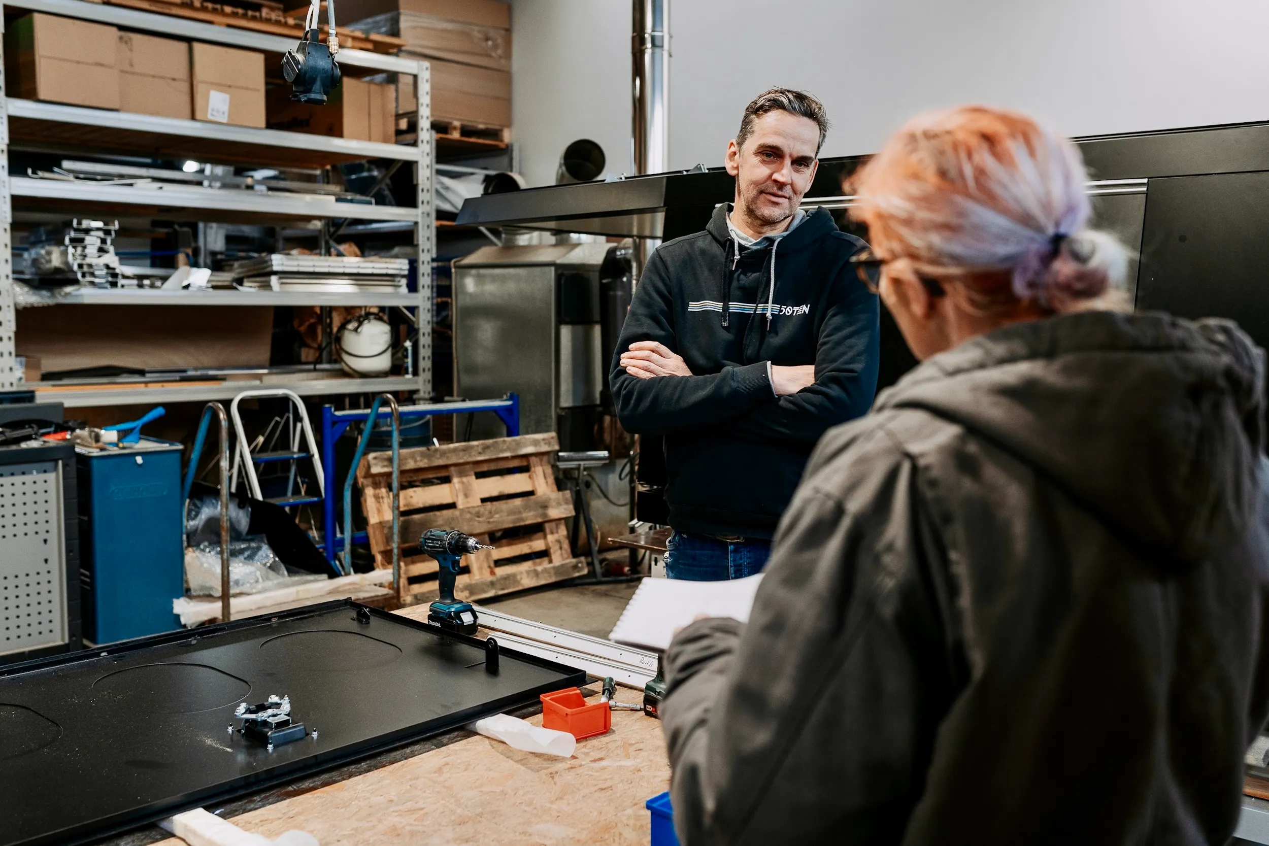 Zwei Personen unterhalten sich in einer Werkstatt mit Regalen, Werkzeug und einer Werkbank mit einem Bohrer und Teilen.