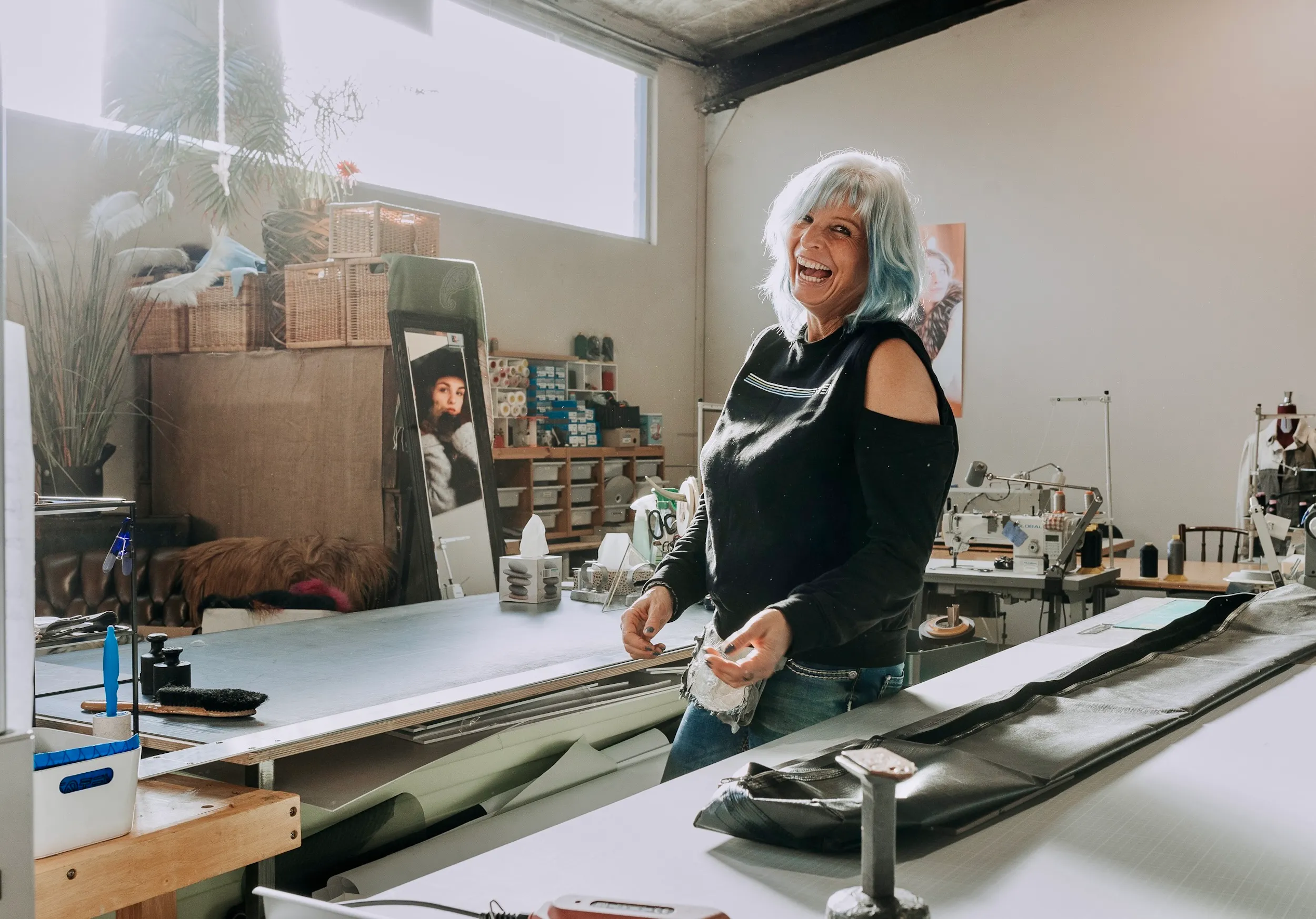 Frau mit blauen Haaren lacht in einem Atelier mit Nähmaschinen und Stoffen.