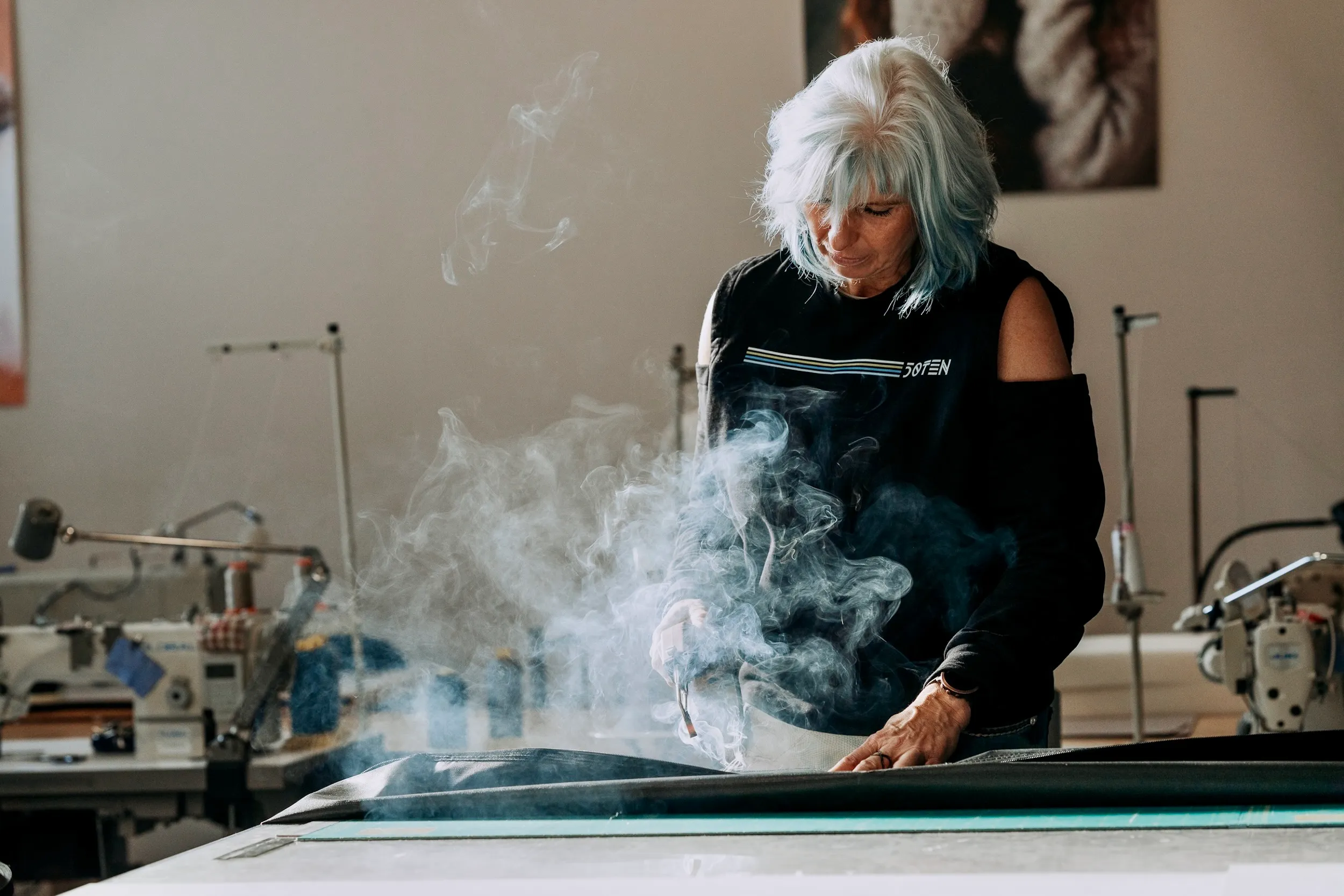 Frau mit weiß-blauen Haaren verwendet ein Bügeleisen, das Dampf über einen Stoff auf einem großen Tisch abgibt, in einer Werkstatt mit Nähausrüstung im Hintergrund.