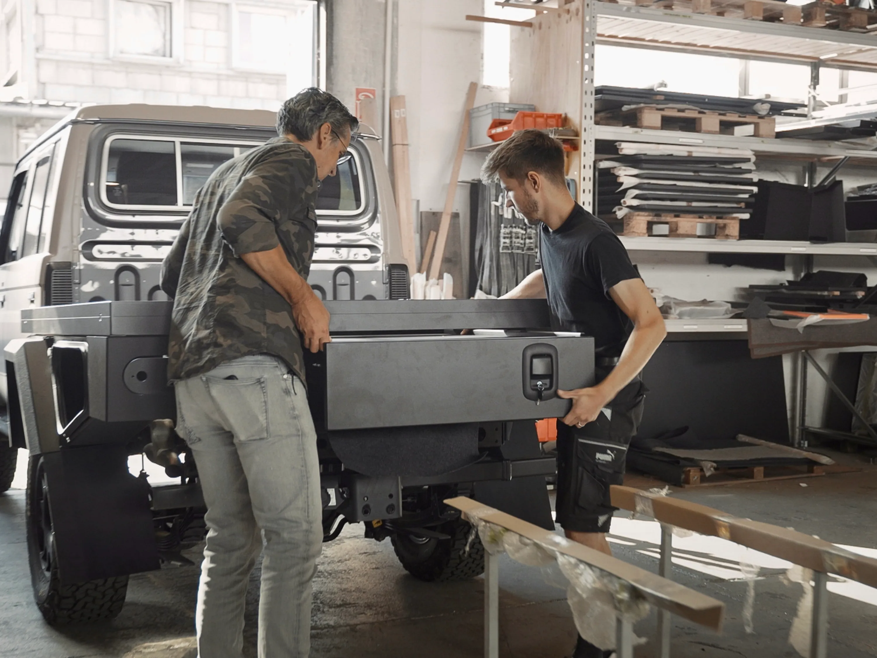 Zwei Männer montieren eine schwarze Lagerbox auf der Ladefläche eines Pick-up-Trucks in einer Werkstatt.