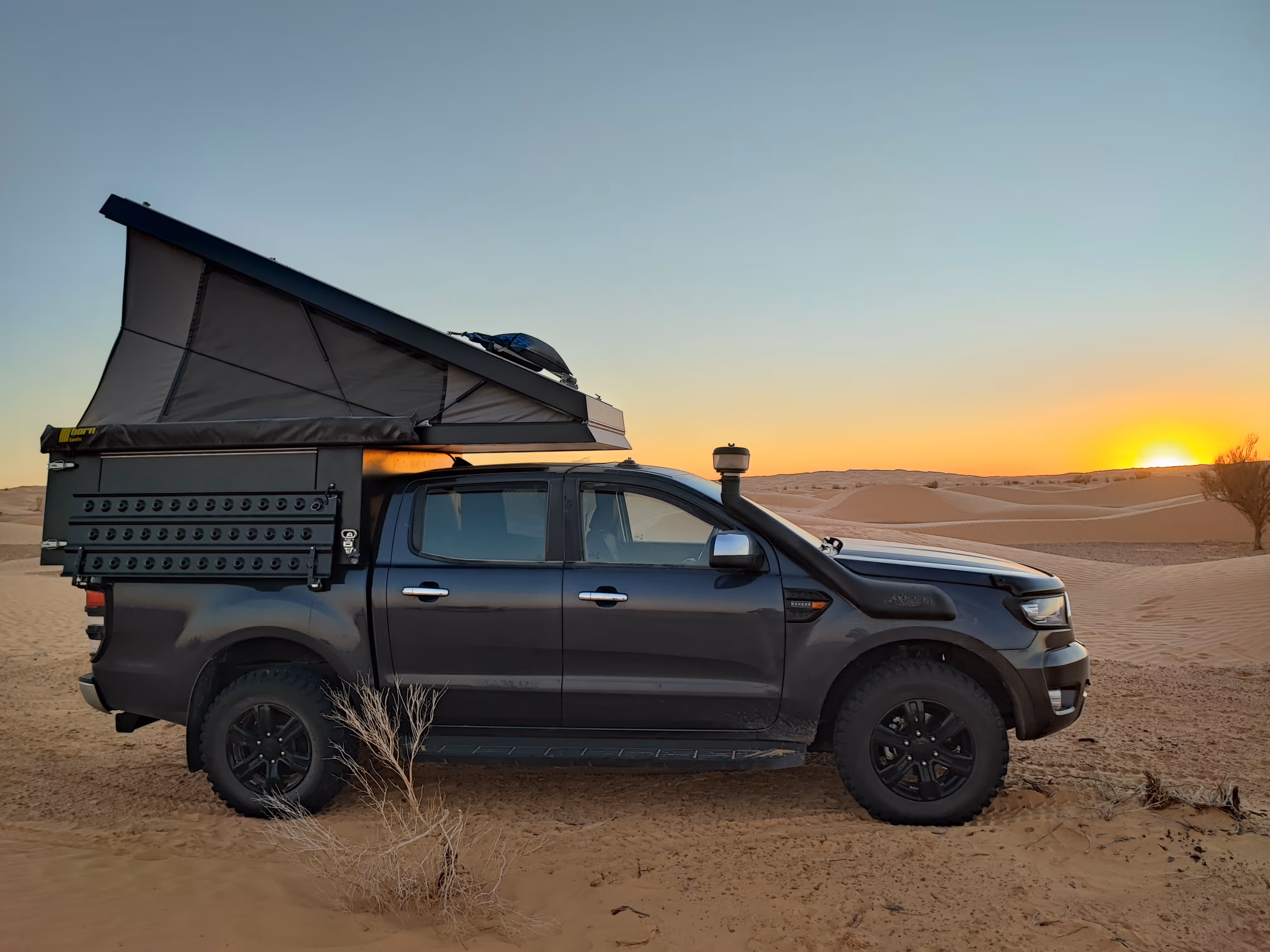 Schwarzer Pick-up mit aufgestelltem Dachzelt im Wüstensand bei Sonnenuntergang.