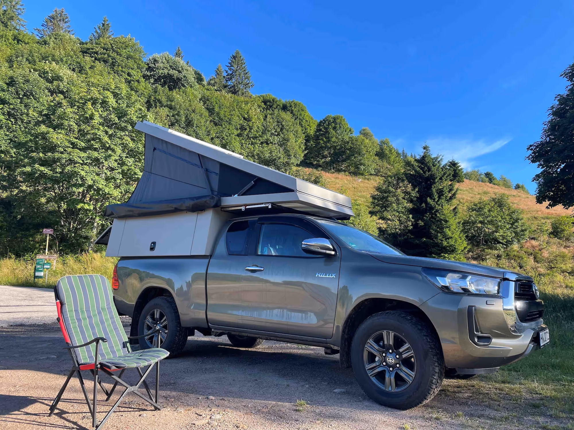 Grauer Toyota Hilux Pick-up mit aufgeklapptem Dachzelt und einem grünen Klappstuhl davor auf einem Parkplatz mit Wald im Hintergrund.