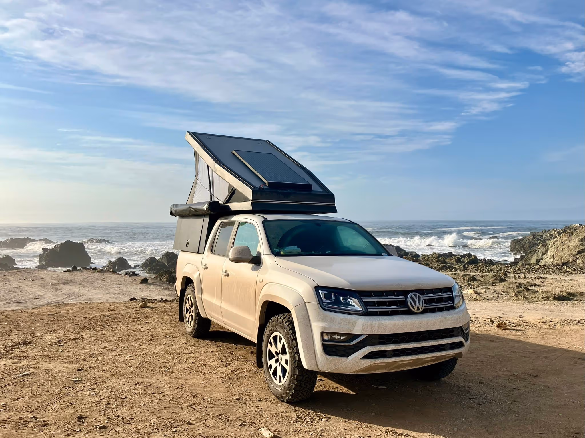 Beiger Volkswagen-Pick-up mit aufgeklapptem Dachzelt und Solarpanel am Meer auf felsigem Strand.