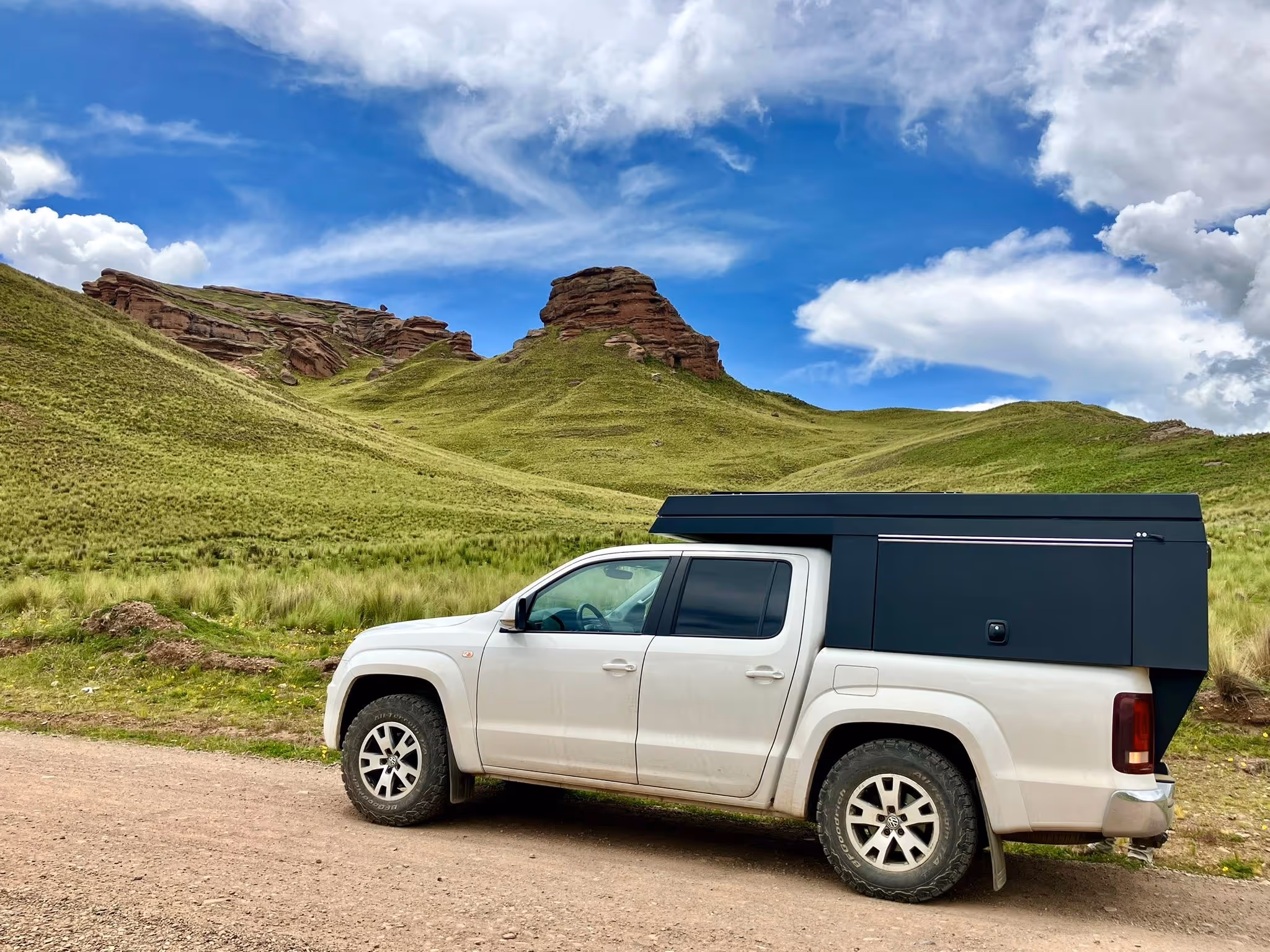 Weißer Pick-up mit schwarzer Aufbaugkabine auf einem Schotterweg vor grünen Hügeln und blauem Himmel mit Wolken.