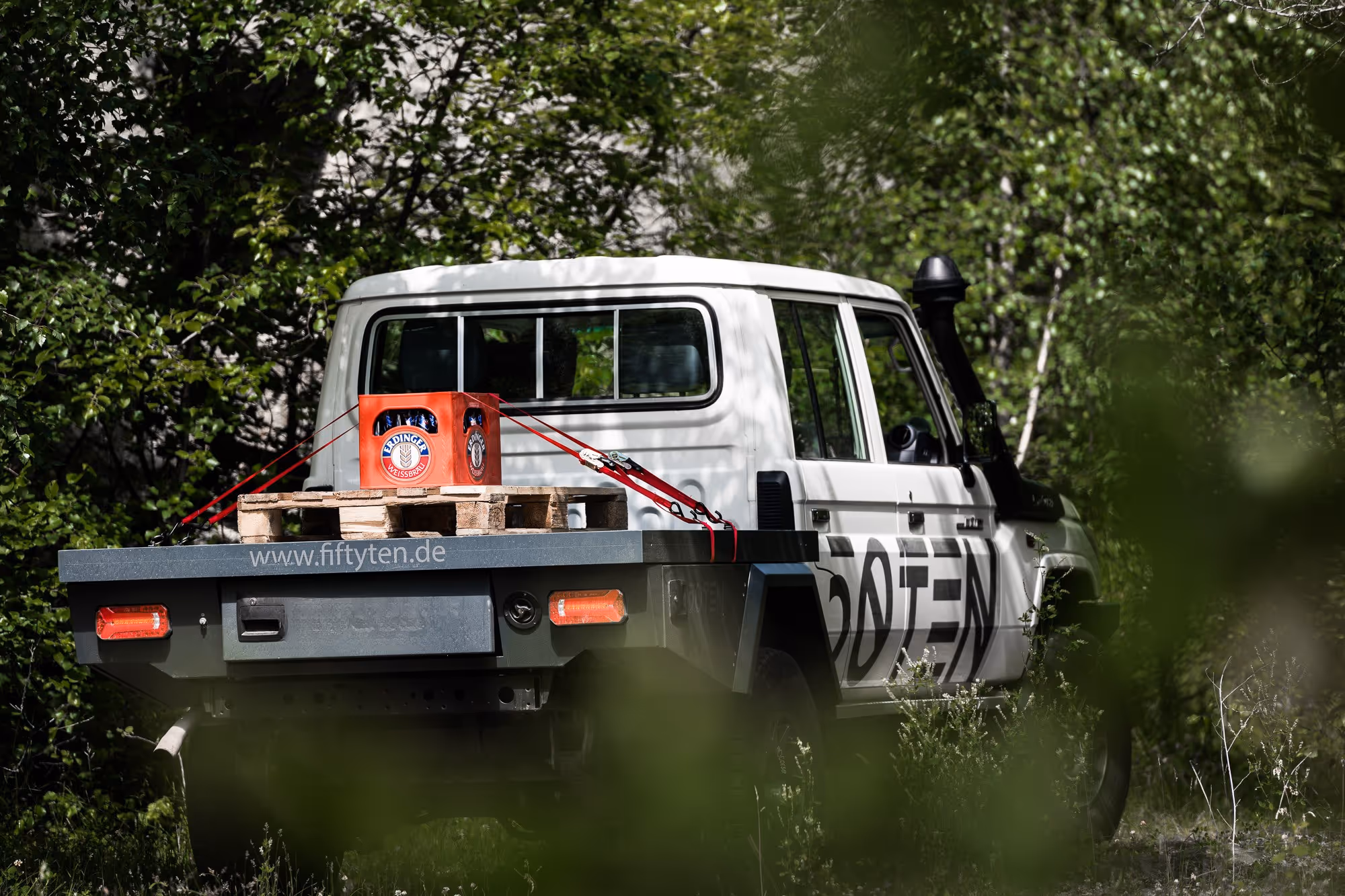 Weißer Geländewagen mit flacher Ladefläche, auf der eine Palette mit einem Kasten Erdinger Weißbier mit roten Spanngurten gesichert ist, umgeben von grünem Wald.