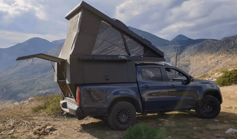 Dunkelblauer Pickup mit aufgestelltem Dachzelt steht auf einem Berg in einer gebirgigen Landschaft unter bewölktem Himmel.