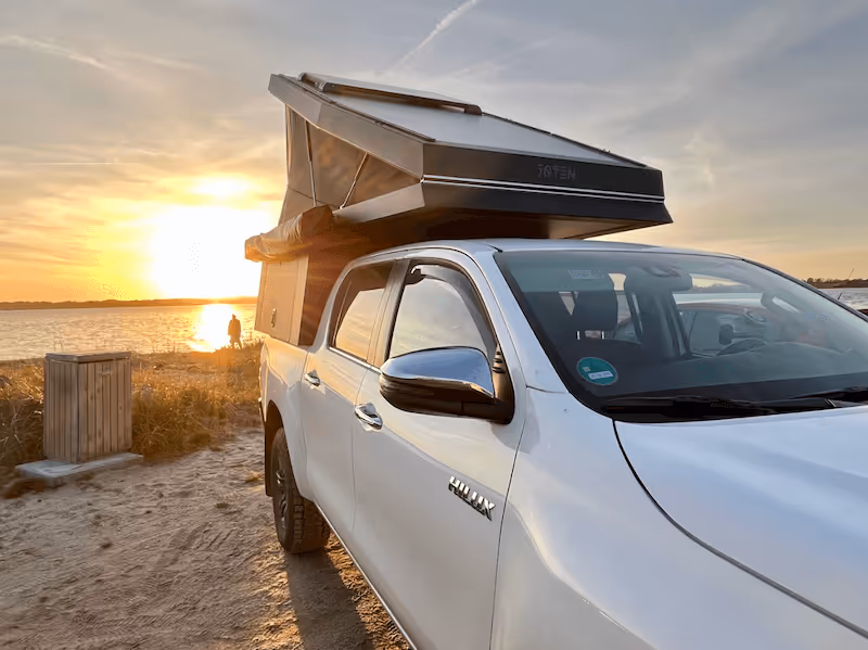 Weißer Pickup mit aufgestelltem Dachzelt bei Sonnenuntergang am Strand neben Wasser.