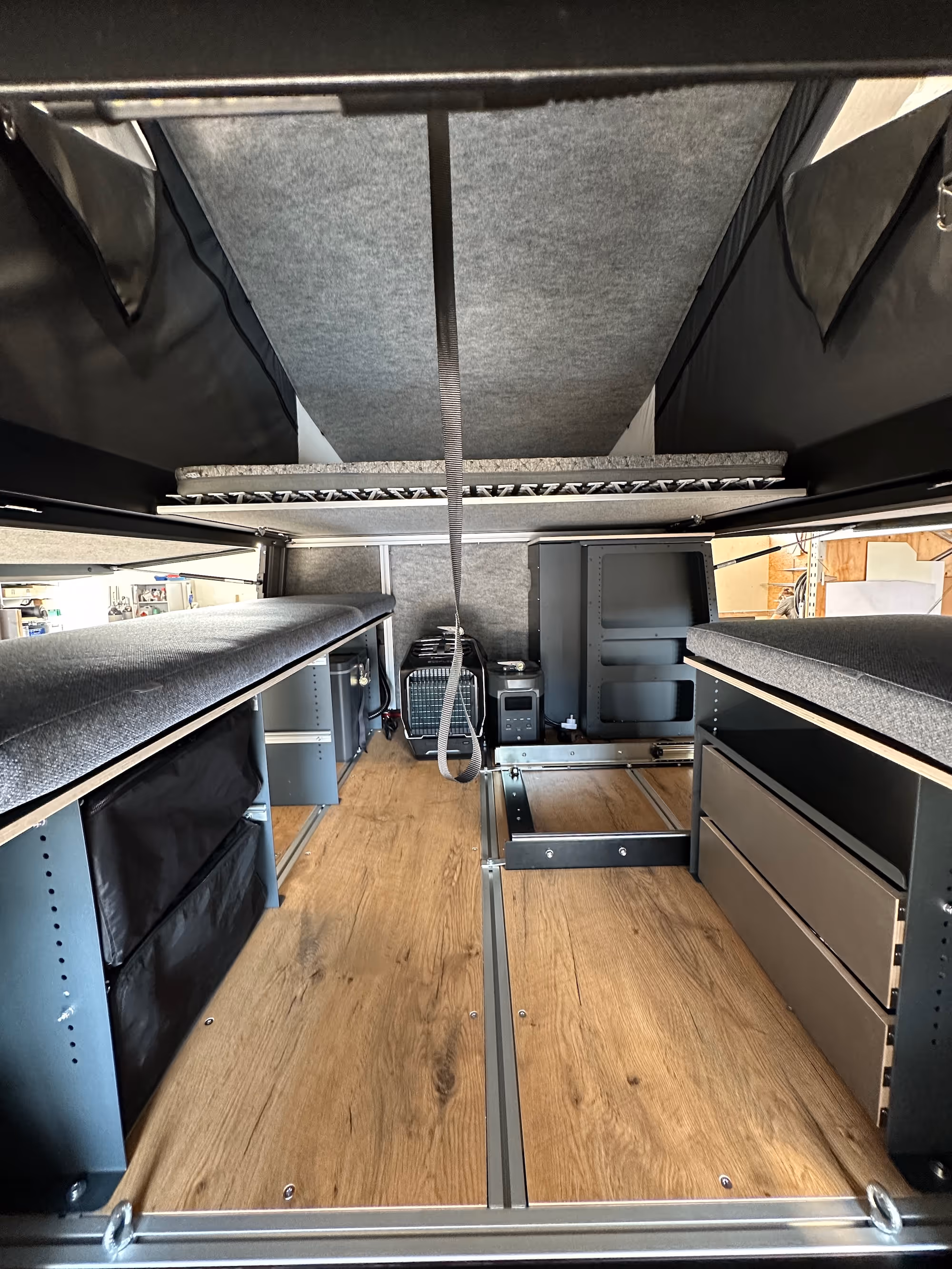 Innenraum eines kompakten Camper-Ausbaus mit Holzboden, gepolsterten Sitzflächen, Schubladen und Regalen.