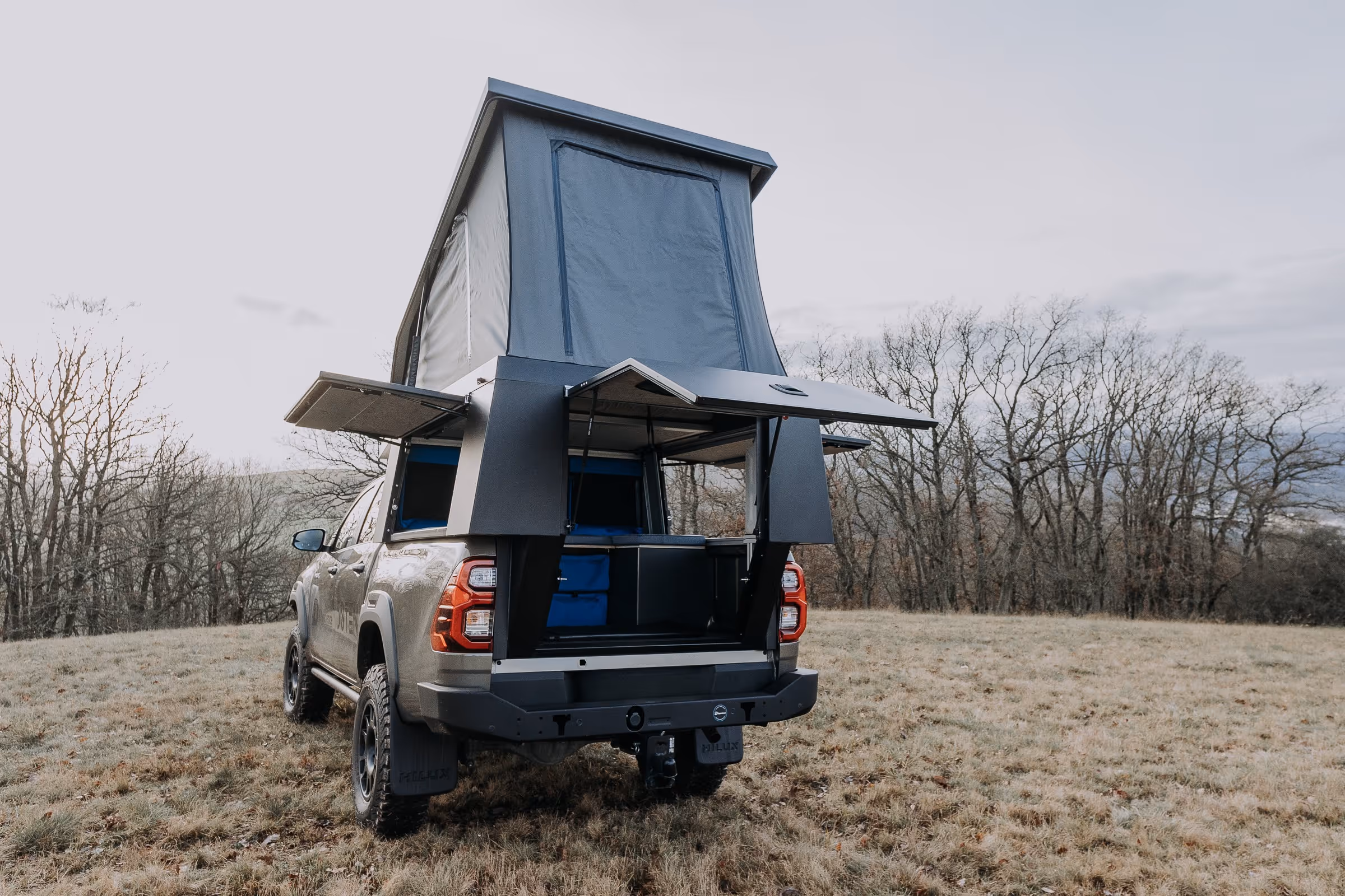 Pick-up-Truck mit ausgeklapptem Camping-Aufbau und Zelt in einer offenen Wiese vor kahlen Bäumen.