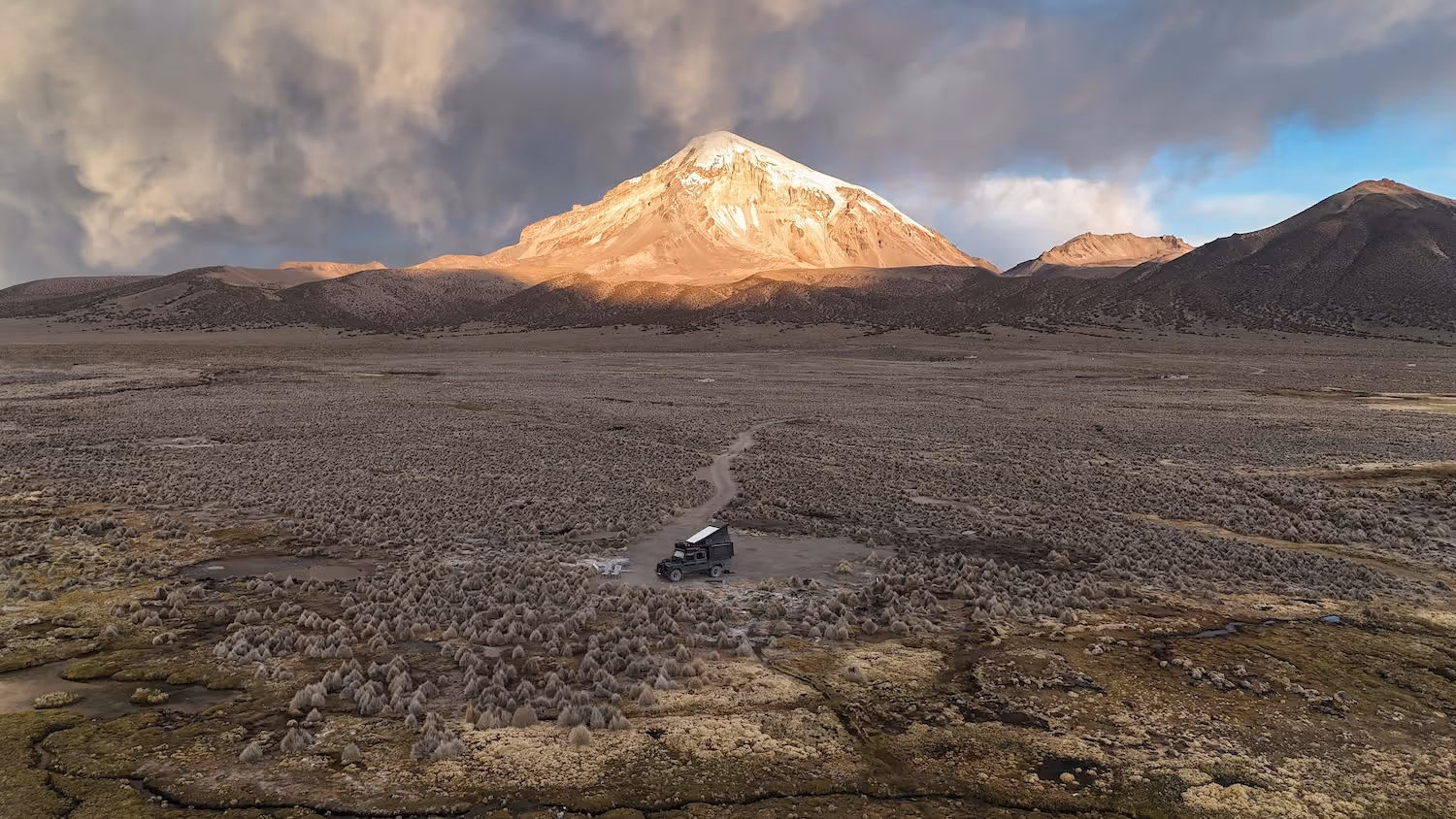 Geländewagen mit aufgestelltem Dachzelt auf karger Landschaft vor einem schneebedeckten Berg unter stürmischem Himmel.