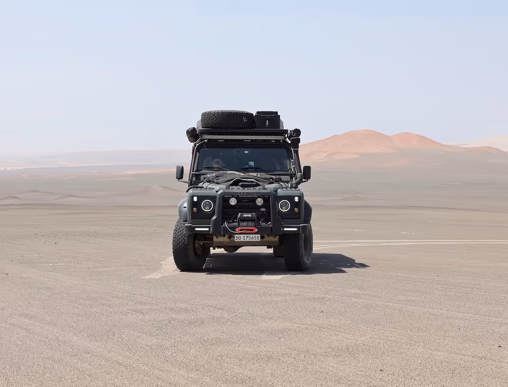 Geländewagen mit Dachgepäck fährt durch eine weite, sandige Wüstenlandschaft mit Bergen im Hintergrund.