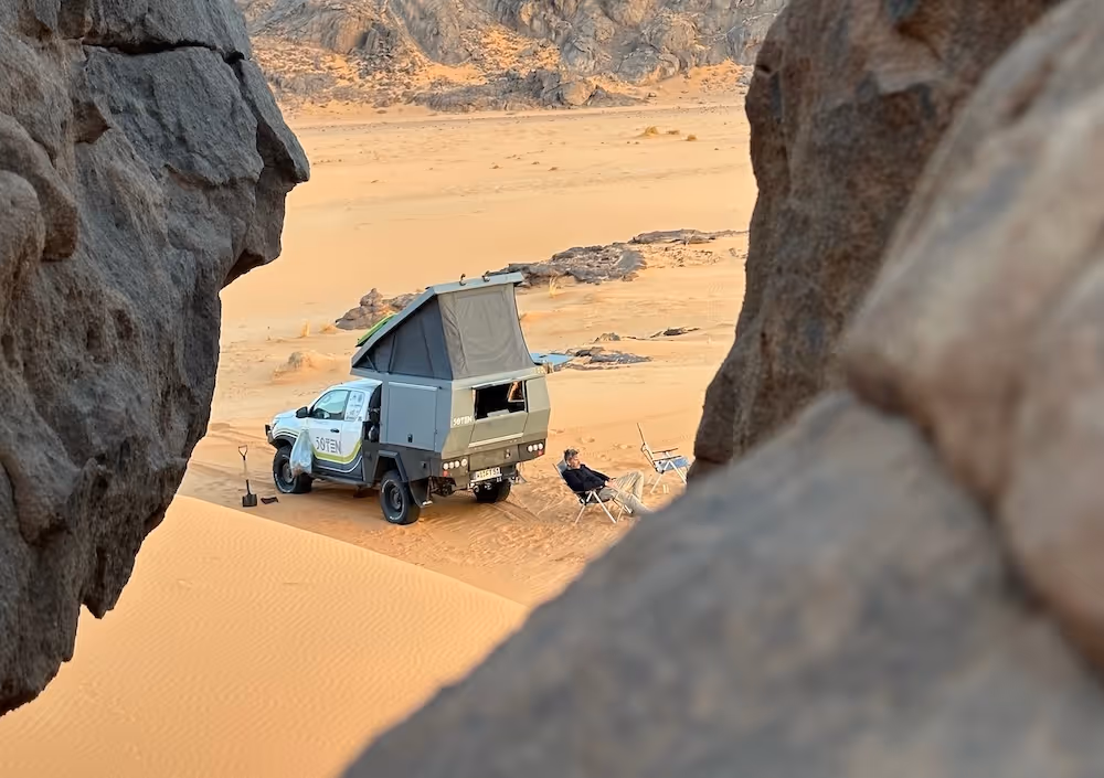 Offroad-Camperfahrzeug mit aufgeklapptem Dach in einer Wüstenlandschaft, daneben zwei Personen in Klappstühlen.