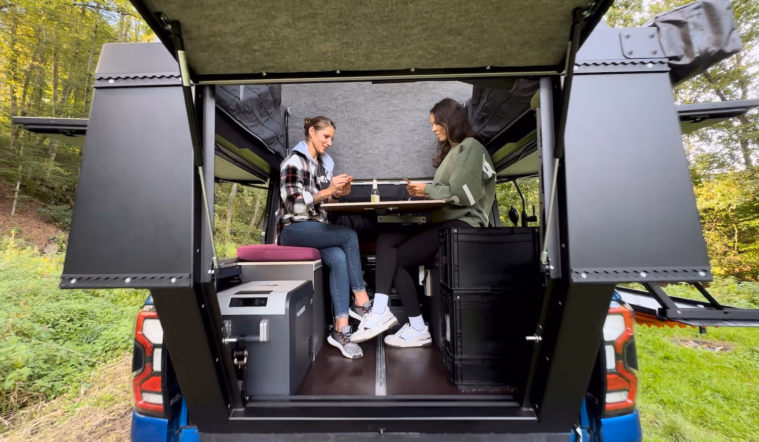 Zwei Frauen sitzen in einem kompakten Wohnaufbau im Heck eines Pick-ups und spielen Karten am kleinen Tisch.