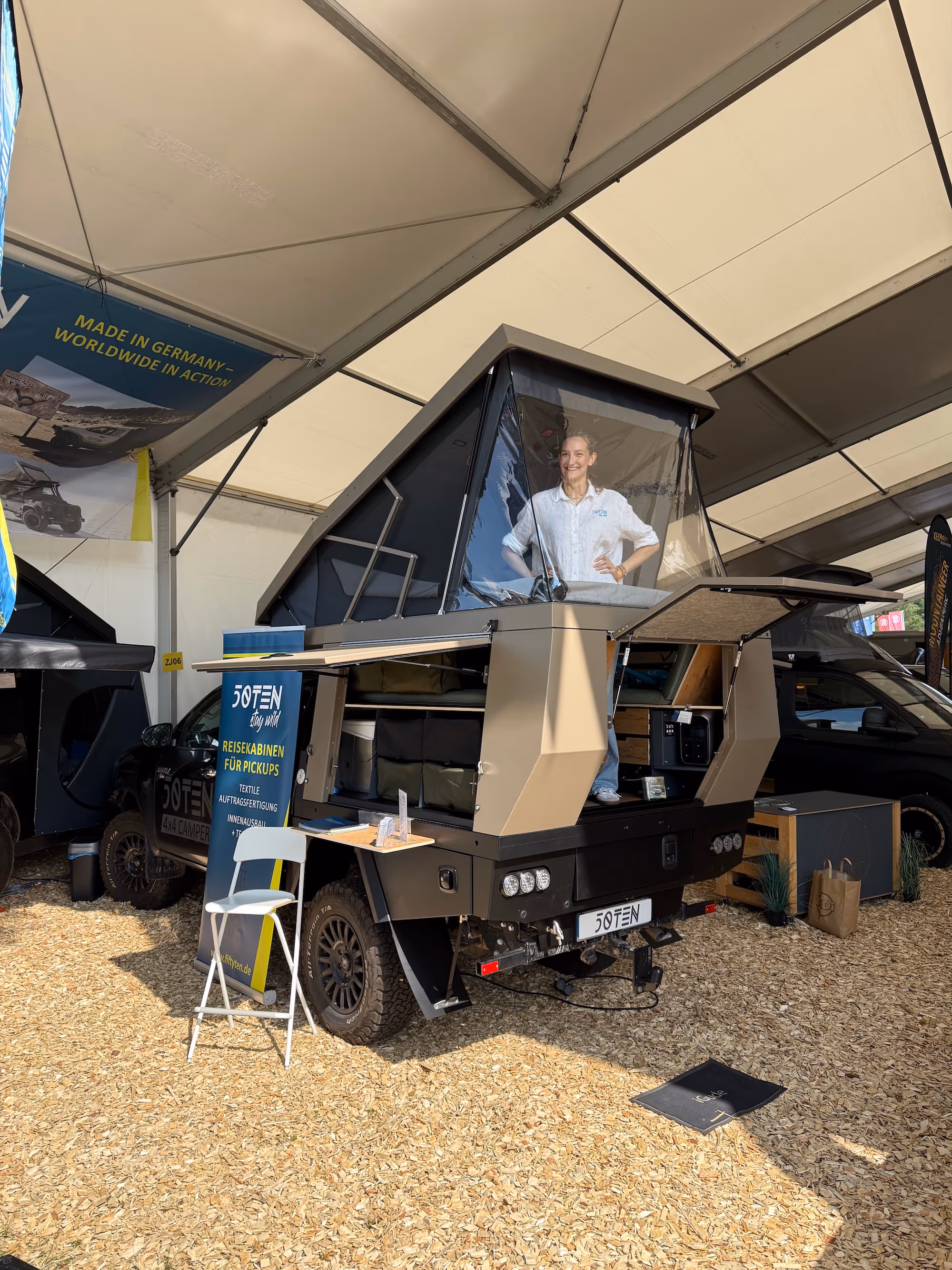 Frau steht in einer geöffneten Übernachtungskabine eines Pickups auf einer Messe unter einem Zelt.