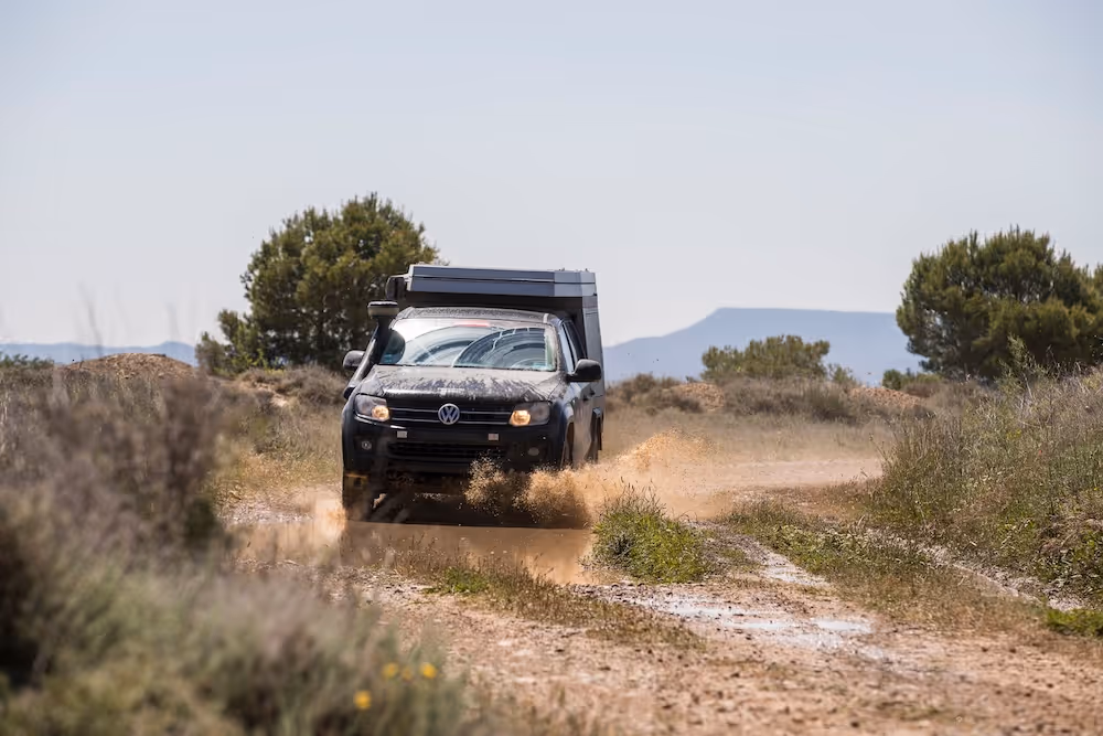 Schwarzer Volkswagen-Pickup fährt auf einer staubigen Landstraße, umgeben von Büschen und Bäumen.