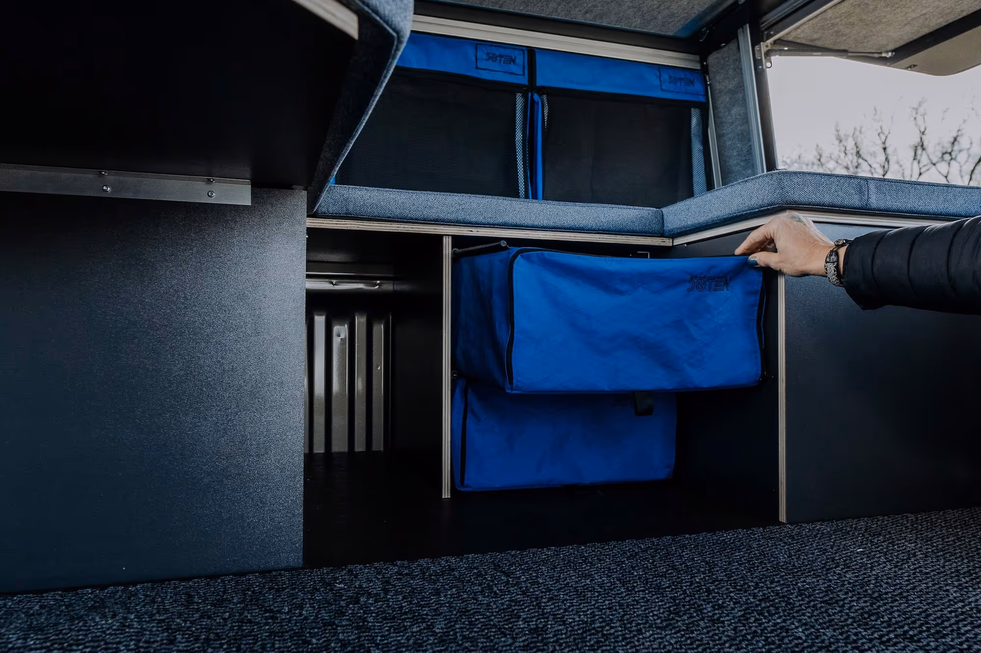 Eine Hand öffnet einen blauen Stoffbehälter in einem modernen Camper-Ausbauschrank mit weiteren blauen Aufbewahrungsboxen.