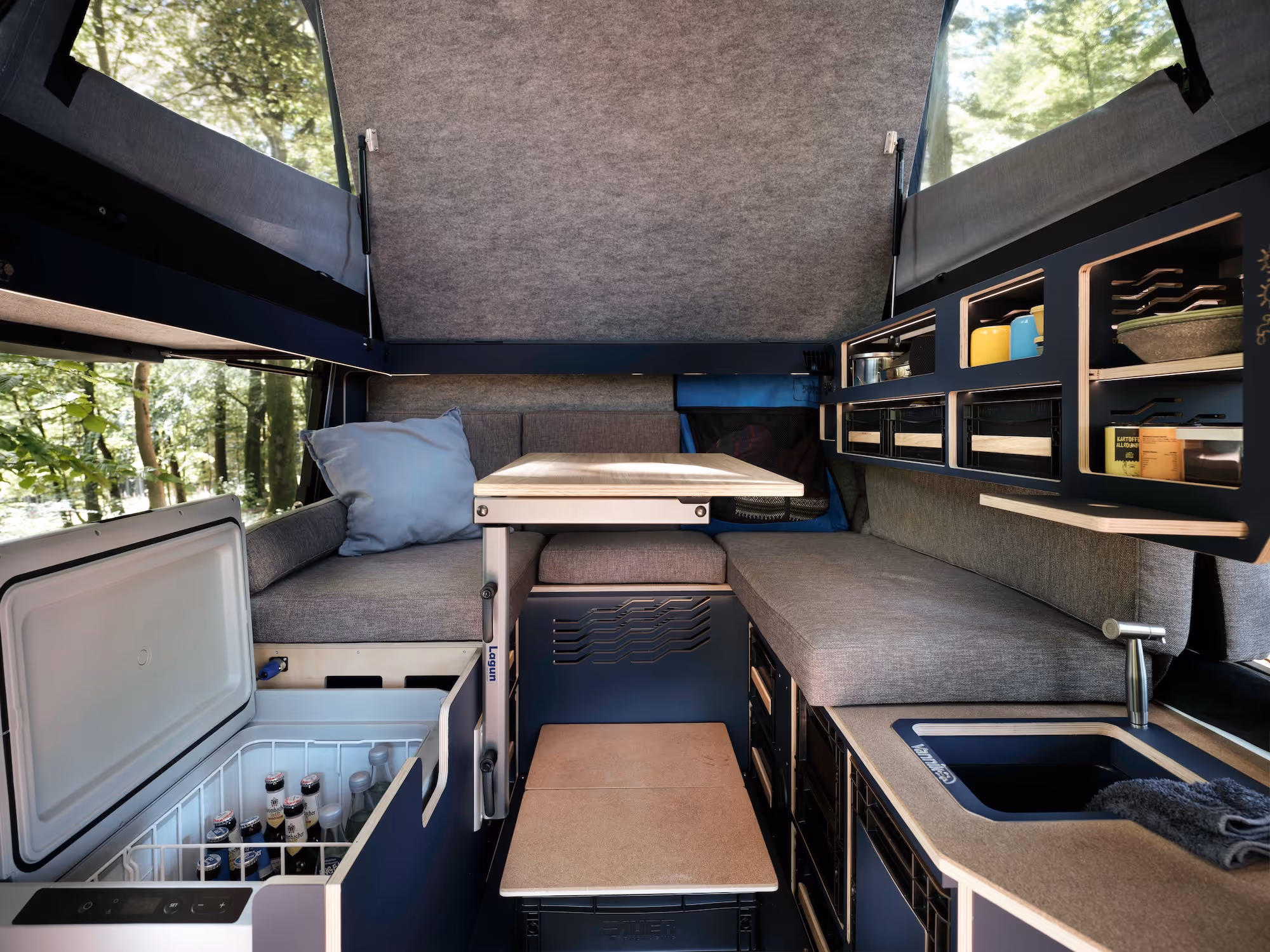 Innenansicht eines kompakt gestalteten Camping-Van mit Sitzbank, Tischen, offener Kühlbox, Waschbecken und Regalen mit Küchenutensilien.
