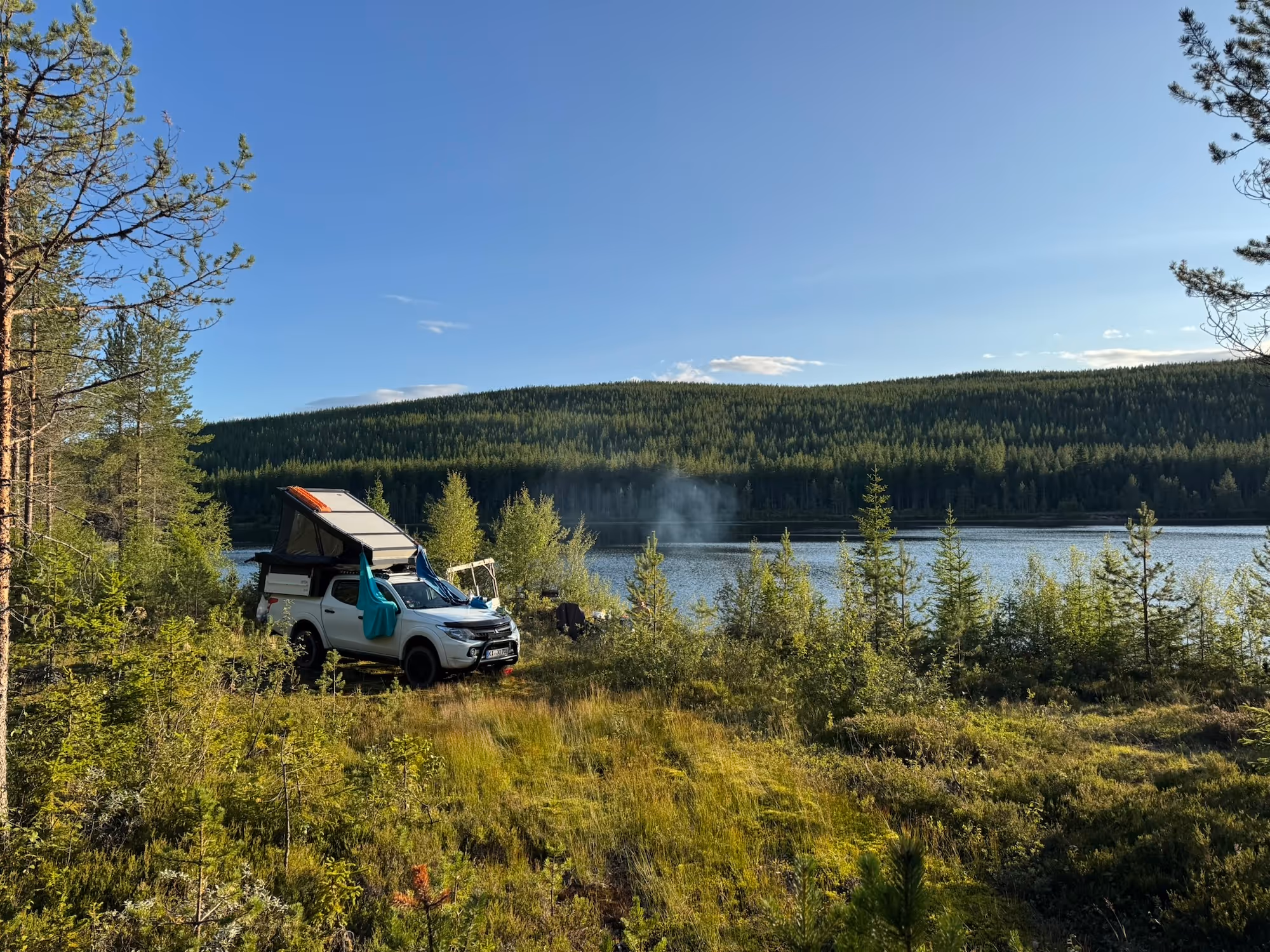 Weißes Camperfahrzeug mit ausgeklapptem Dachzelt, das an einem bewaldeten See bei sonnigem Himmel geparkt ist.