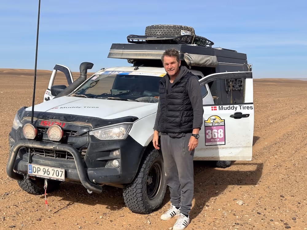 Mann steht vor einem weißen Isuzu-Geländewagen mit Muddy Tires Aufkleber und Dachgepäck, im Hintergrund weite Wüstenlandschaft.