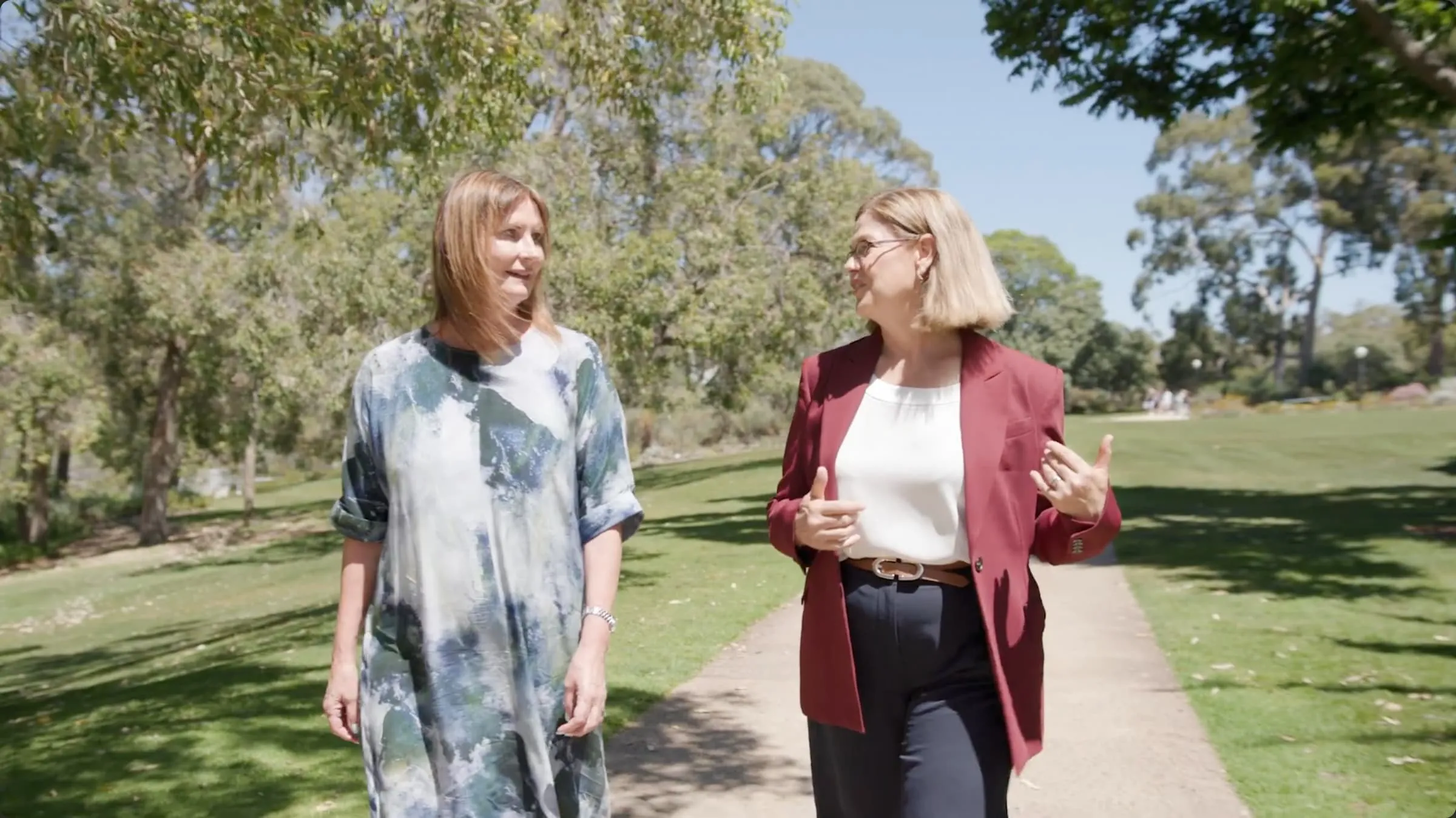 Two women walking and talking on a sunny paved path in a green park with trees and grass.