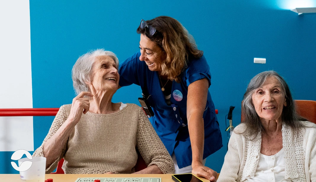 Aide-soignant avec résidentes en salle commune et le boîtier Orikio en fond.