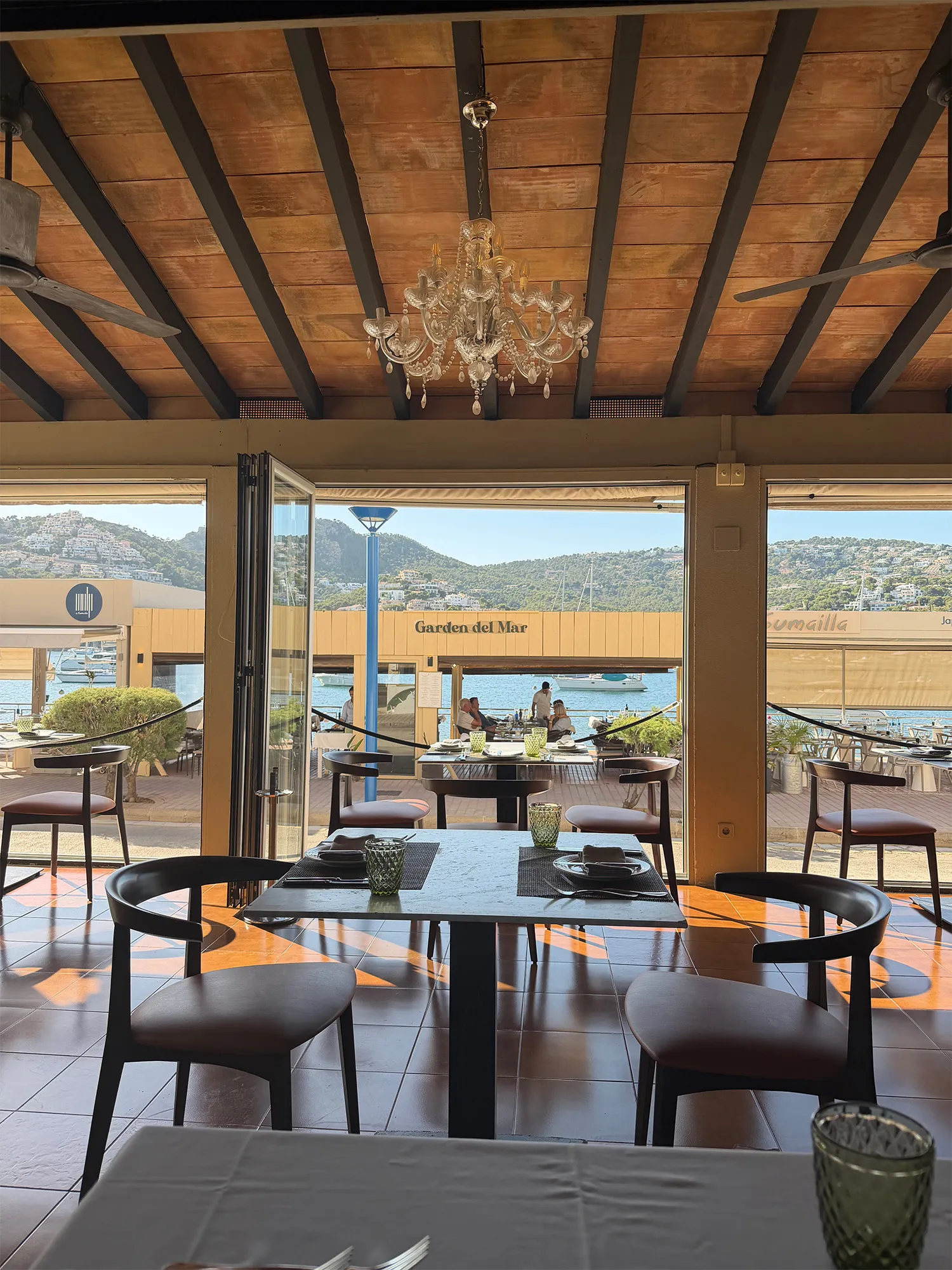 Indoor restaurant seating with tables and chairs under wooden ceiling and chandelier, view of outdoor seating and sea.