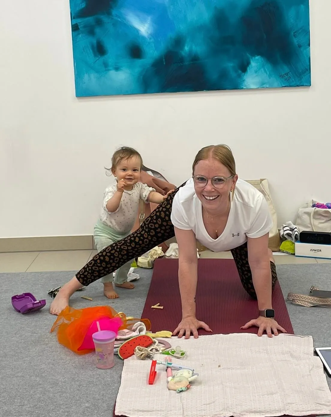Frau praktiziert Mama-Baby-Yoga auf der Matte, Baby spielt im Raum.