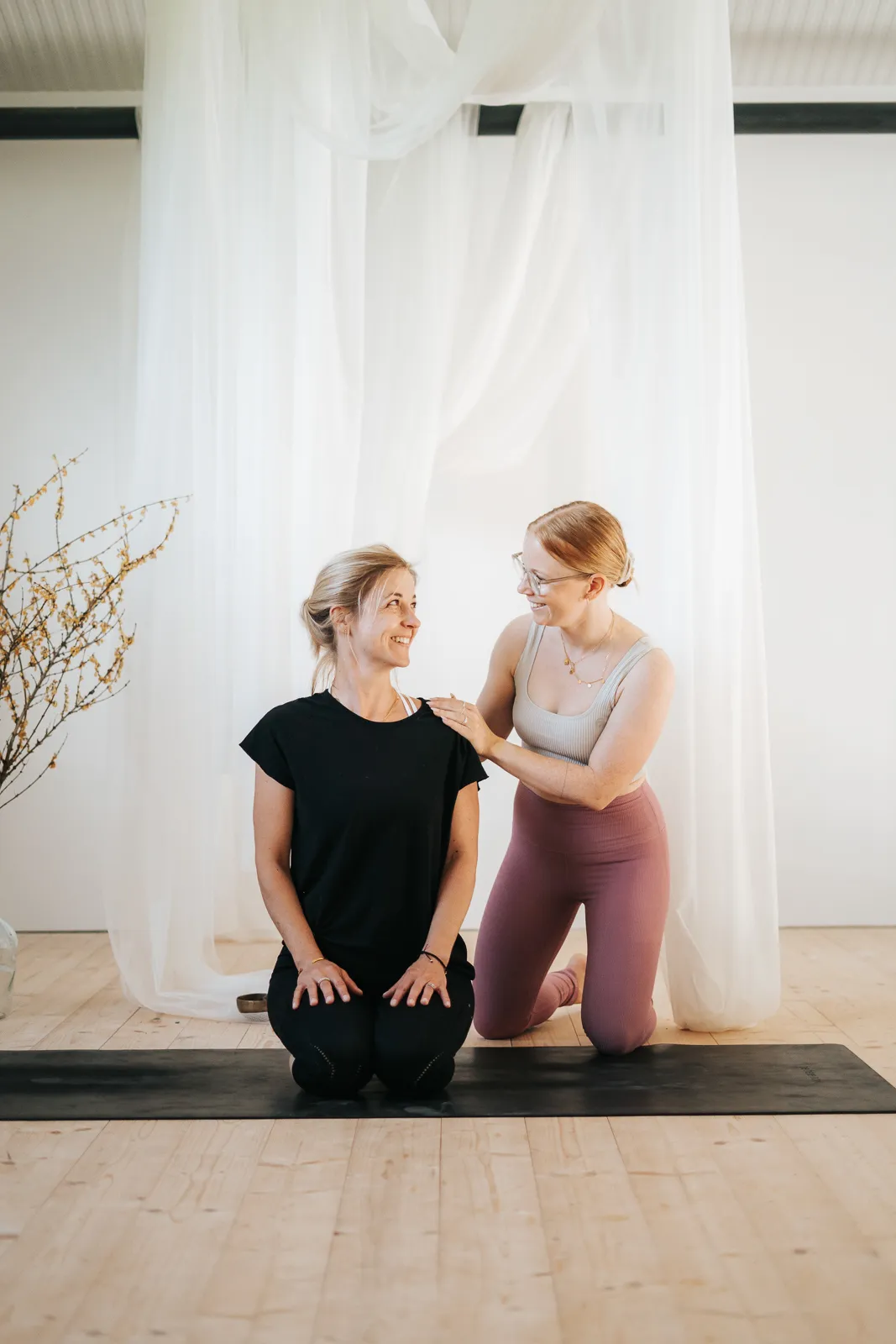 Yogalehrerin gibt persönliche Hilfestellung an Teilnehmerin in kniender Haltung während einer Yoga-Stunde in Niederösterreich.