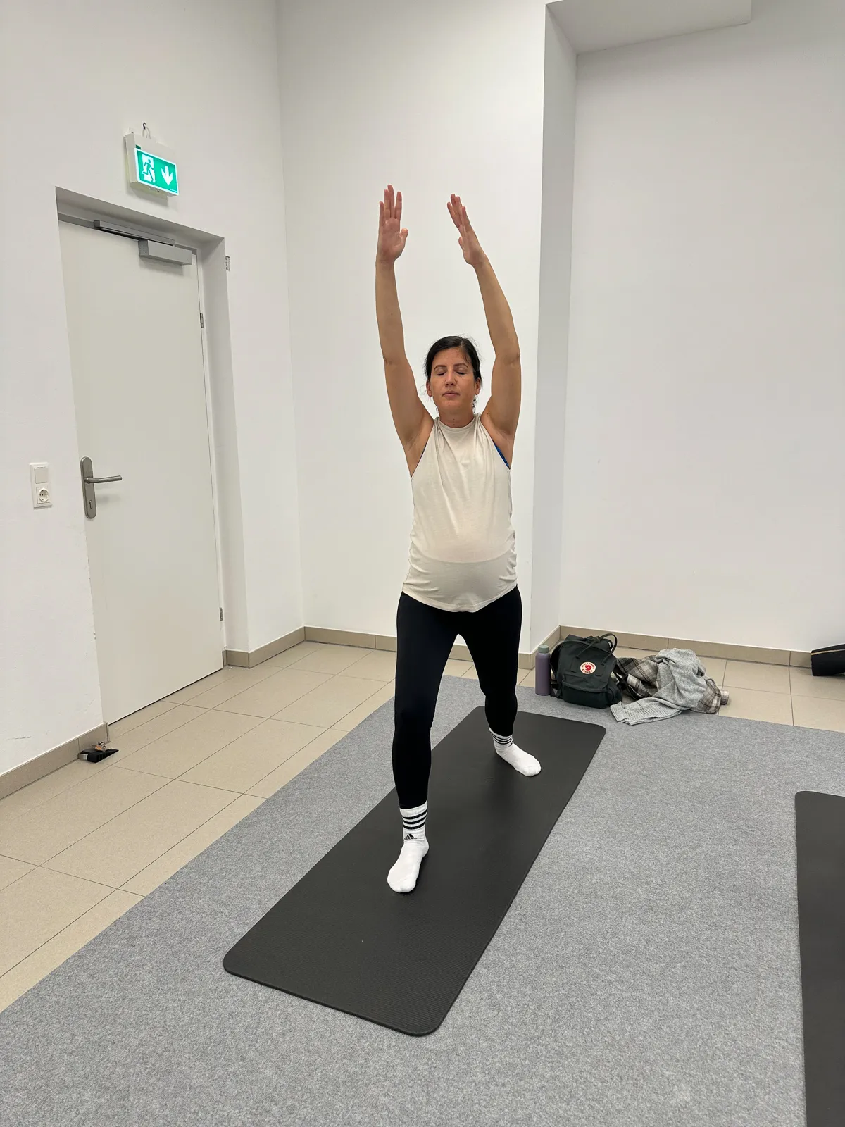 Schwangere Frau steht auf der Yogamatte in der Krieger-Pose mit erhobenen Armen, Teil einer sanften Schwangerschaftsyoga-Praxis.