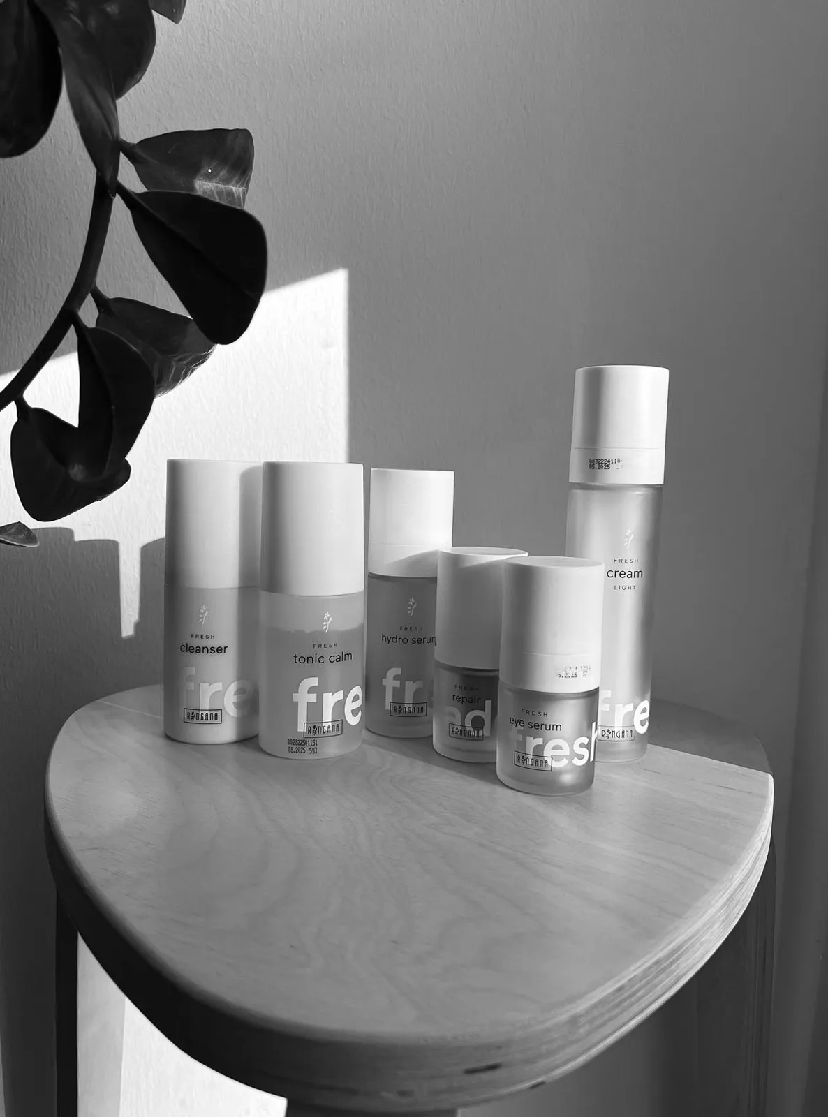 Ringana Frischekosmetik Pflegeprodukte in Schwarz-Weiß aufgenommen, darunter Cleanser, Tonic Calm, Hydro Serum, Repair Serum, Eye Serum und Cream Light auf einem Holzstuhl.
