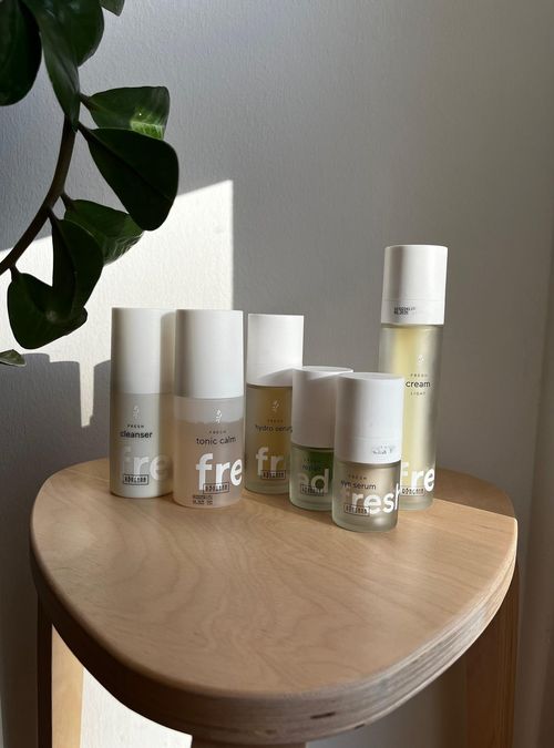 Verschiedene Ringana Frischekosmetik Produkte wie Cleanser, Tonic Calm, Hydro Serum, Repair Serum, Eye Serum und Cream Light auf einem Holzstuhl mit Pflanze im Hintergrund.