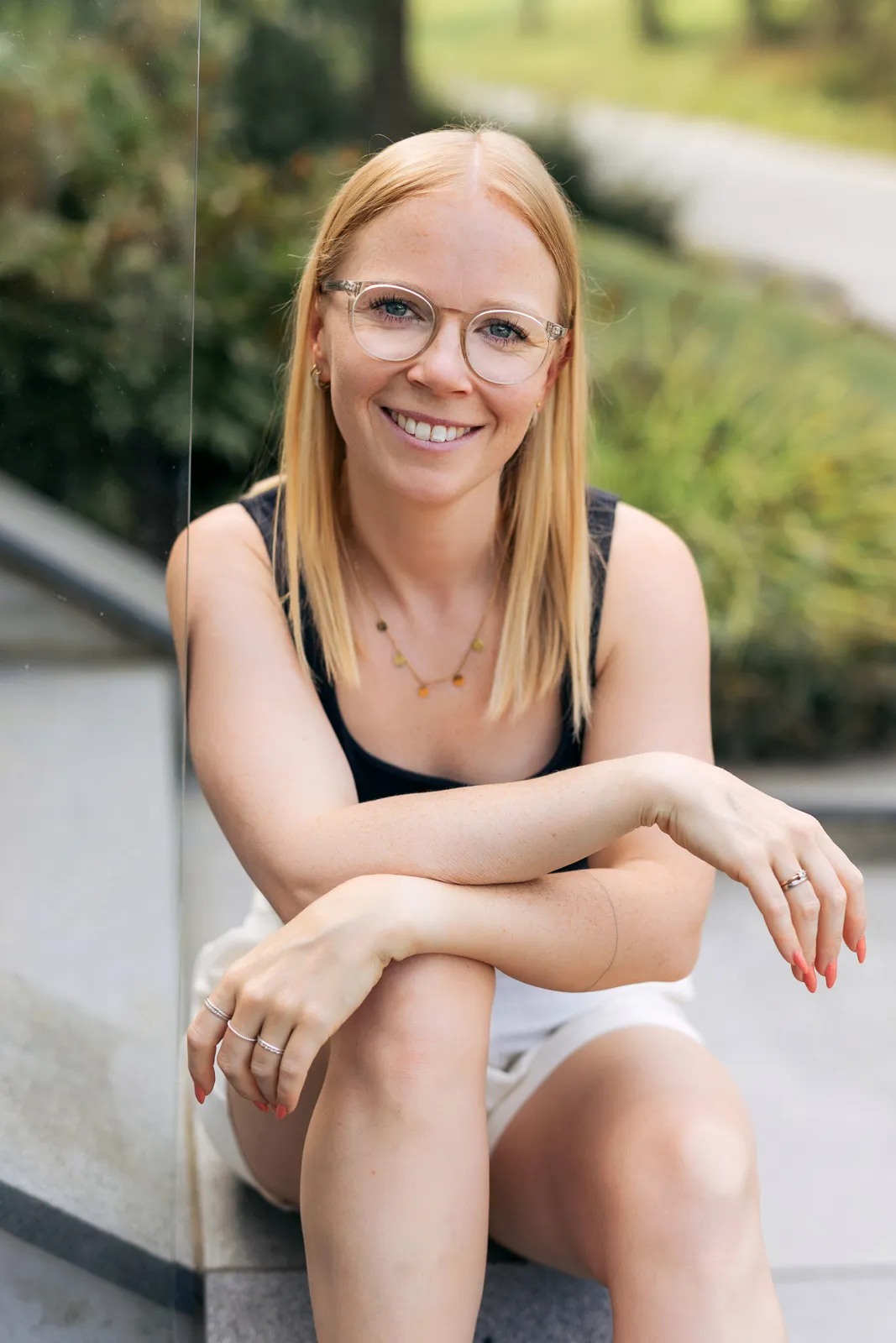 Frau sitzt lächelnd auf Stufen, trägt schwarze Bluse und weiße Shorts, freundliche Ausstrahlung